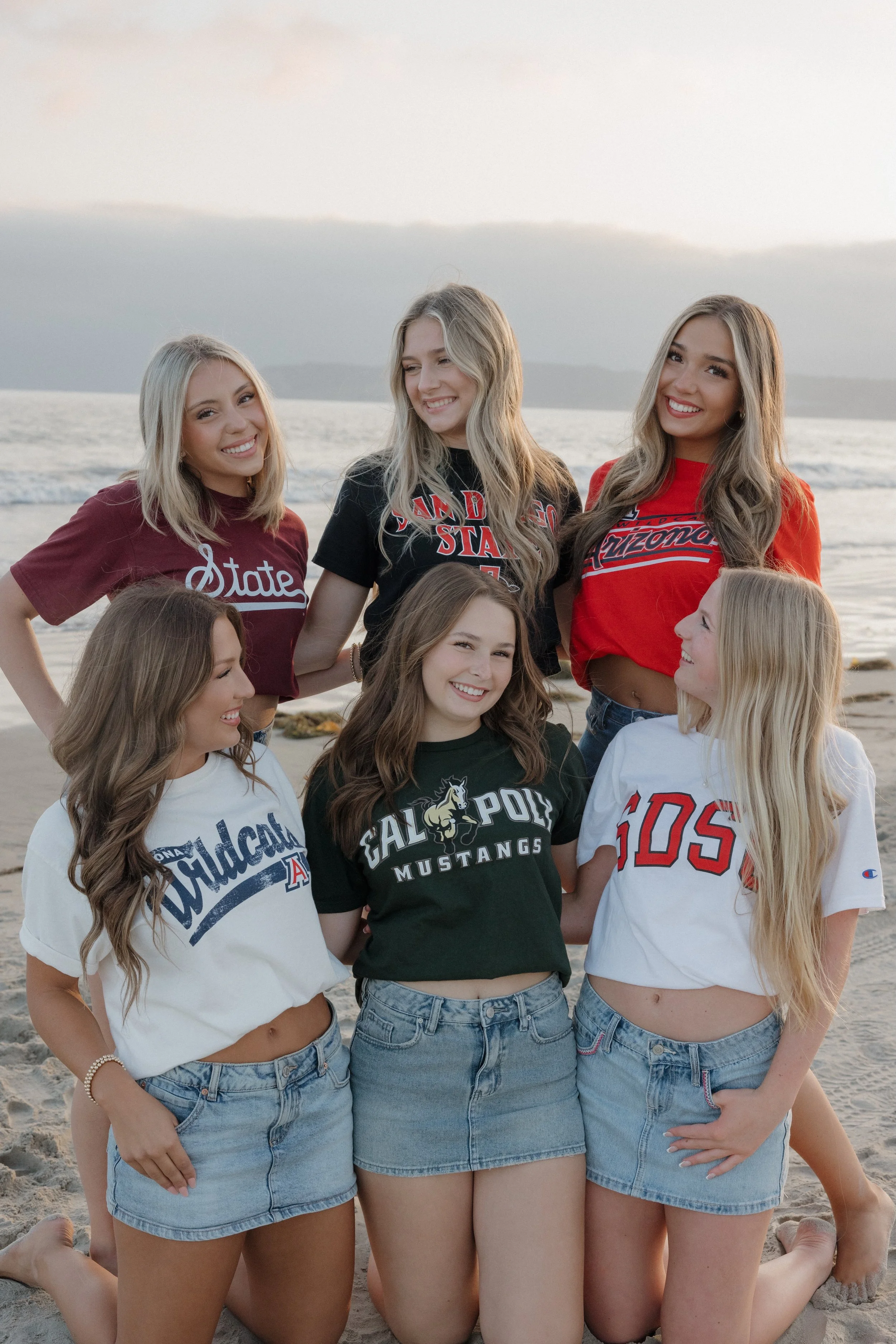 Group of young women on the beach, wearing university or college team shirts, some kneeling and some standing, smiling and looking at each other with the ocean and sky in the background during sunset.