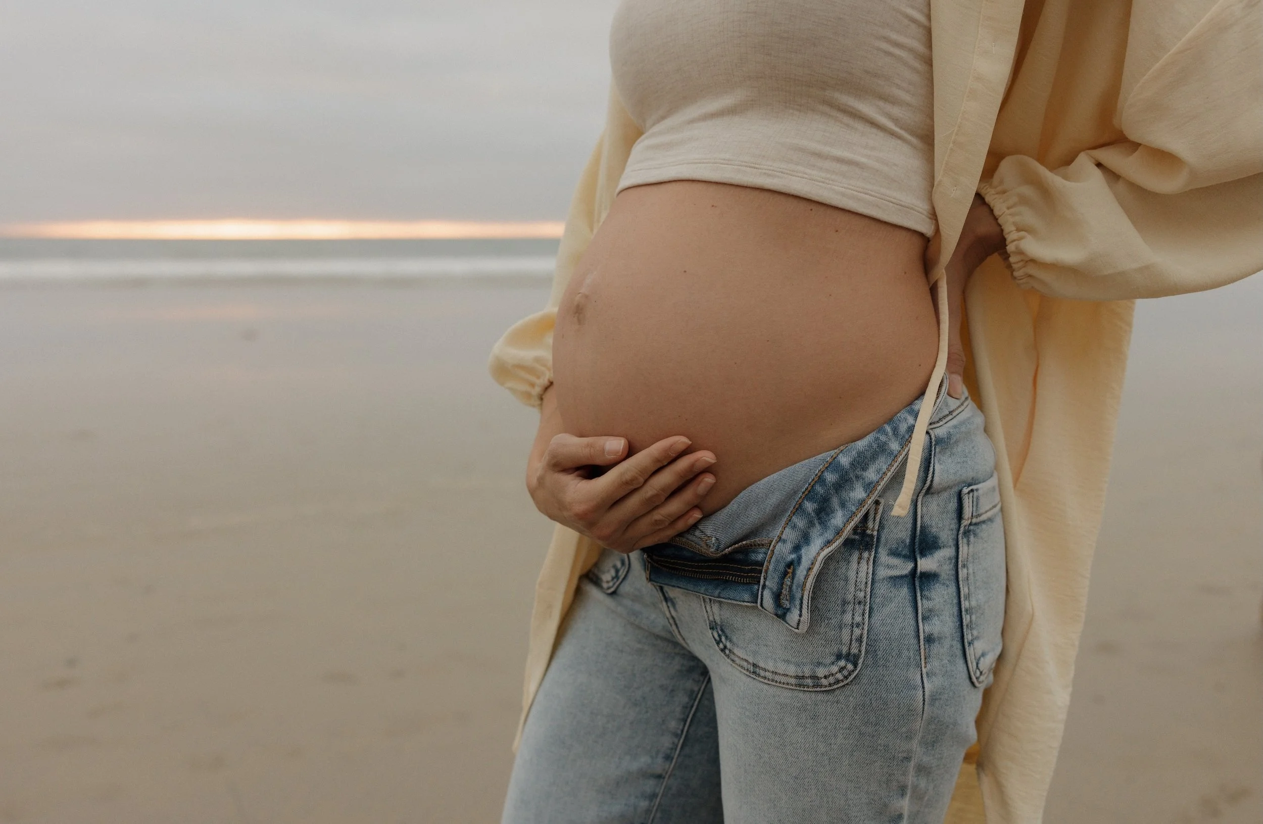 A pregnant woman standing on a beach, wearing a beige jacket, light-colored crop top, and denim shorts, with her hand resting on her belly. The background shows a shoreline and the sky.