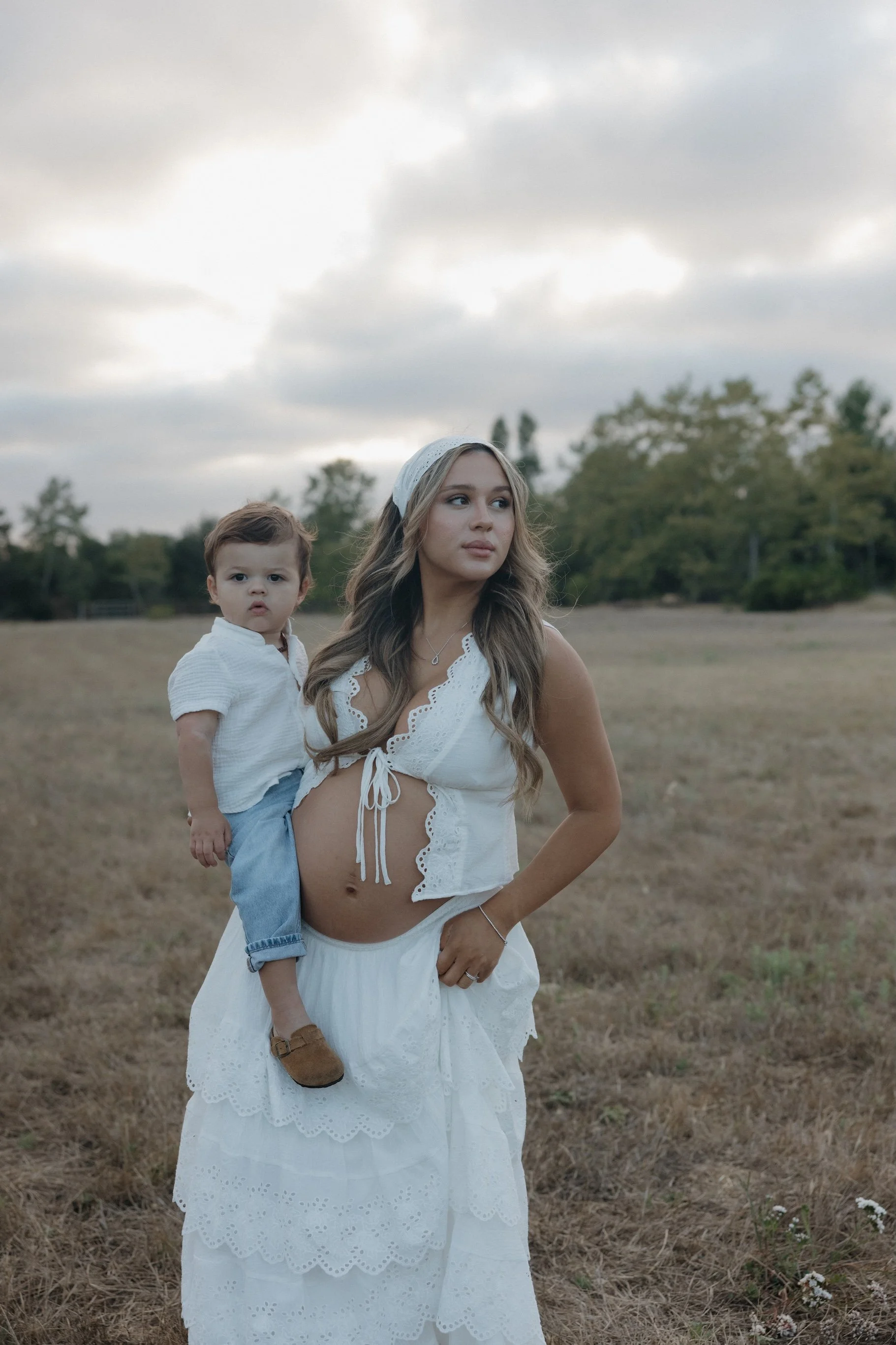 A woman in a white skirt and sleeveless top holding a young boy, both outdoors in a field with trees and gray clouds overhead.