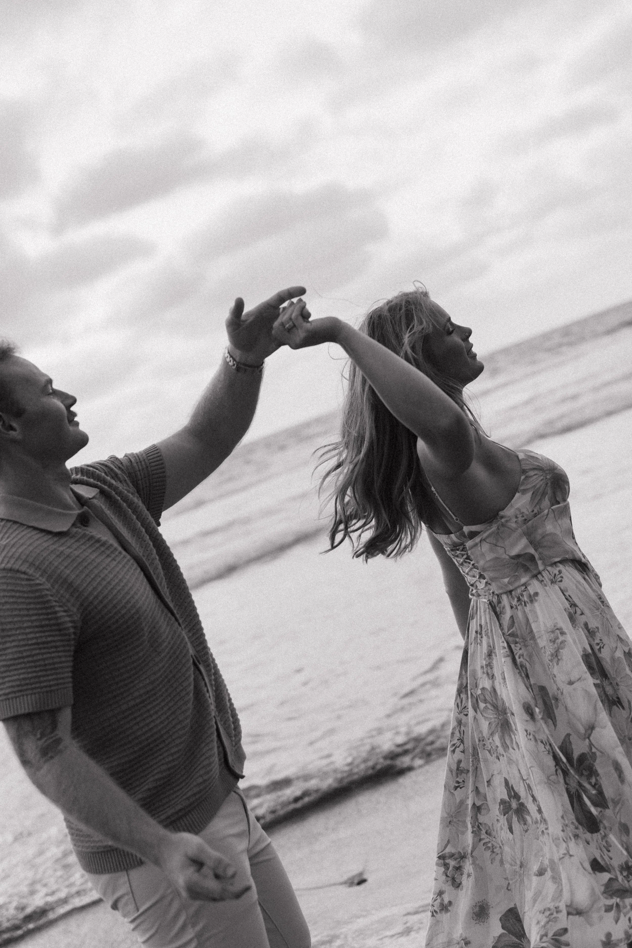 A black and white photo of two women at the beach. One woman is smiling and the other woman, with long hair, is turning her head back and smiling. They are holding hands and the ocean and horizon can be seen in the background.