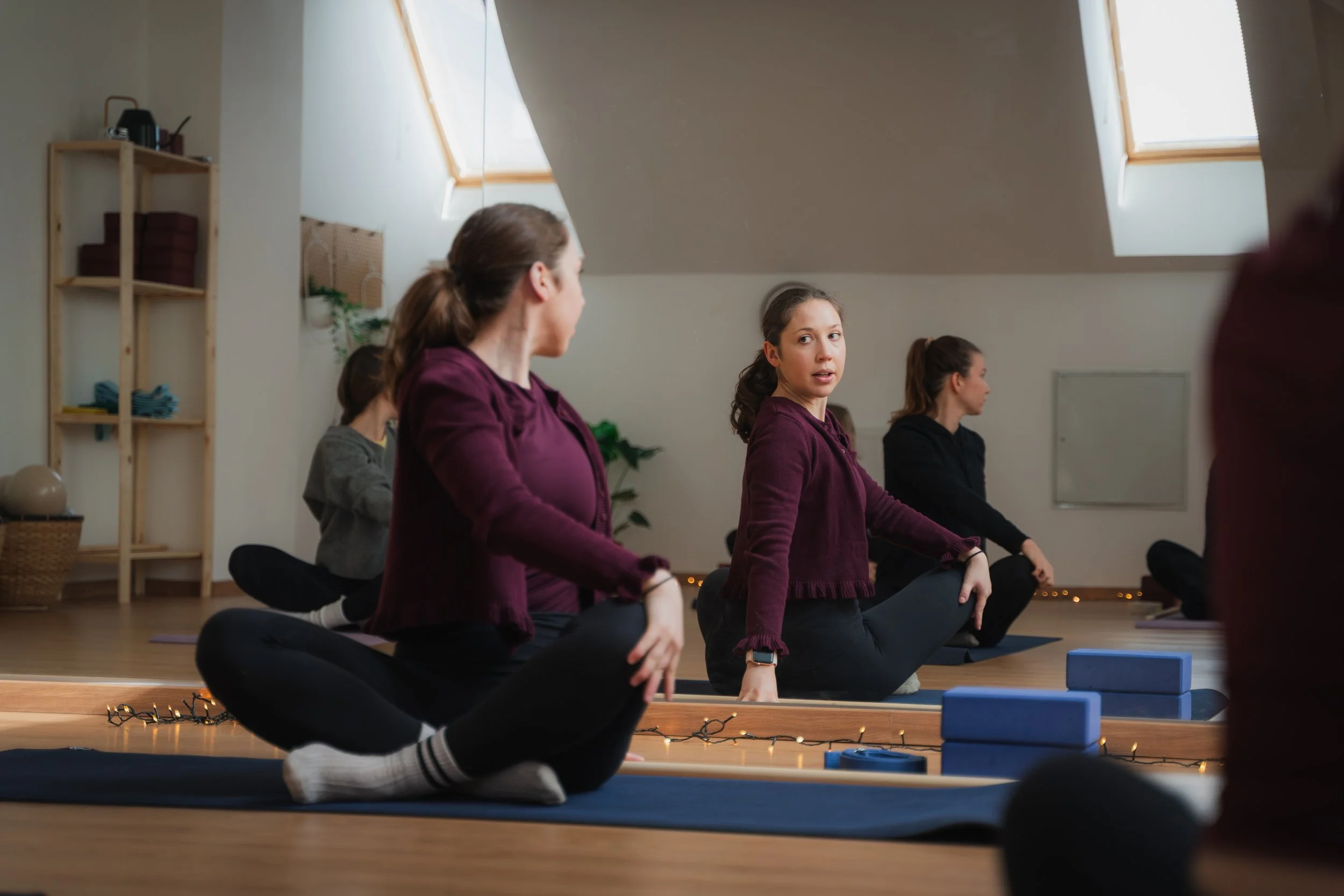Women practicing yoga in a studio with skylight windows, sitting cross-legged on mats, with one woman looking at the instructor.