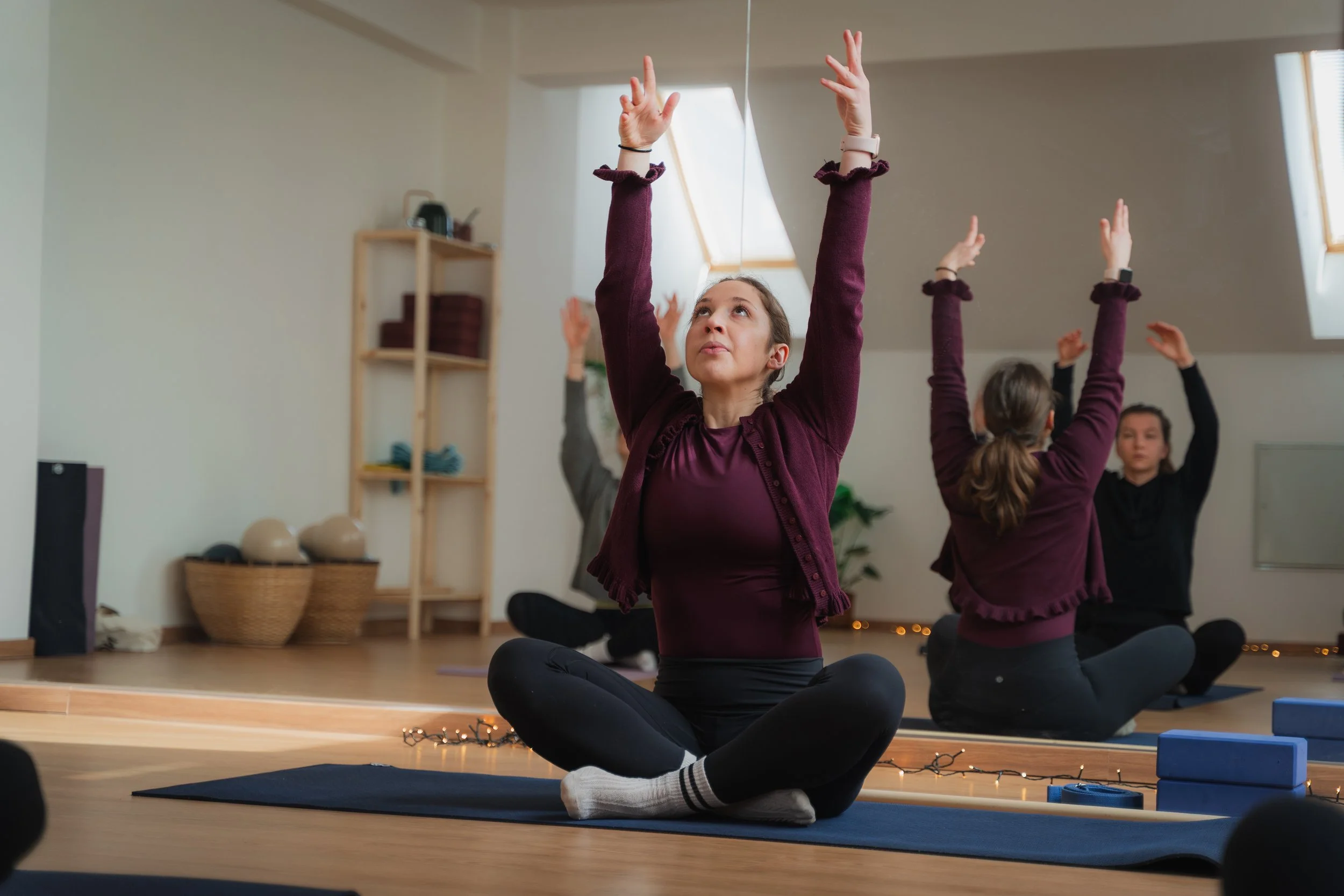 Women participating in a yoga or meditation class, sitting cross-legged on yoga mats with their arms raised, in a well-lit indoor studio.