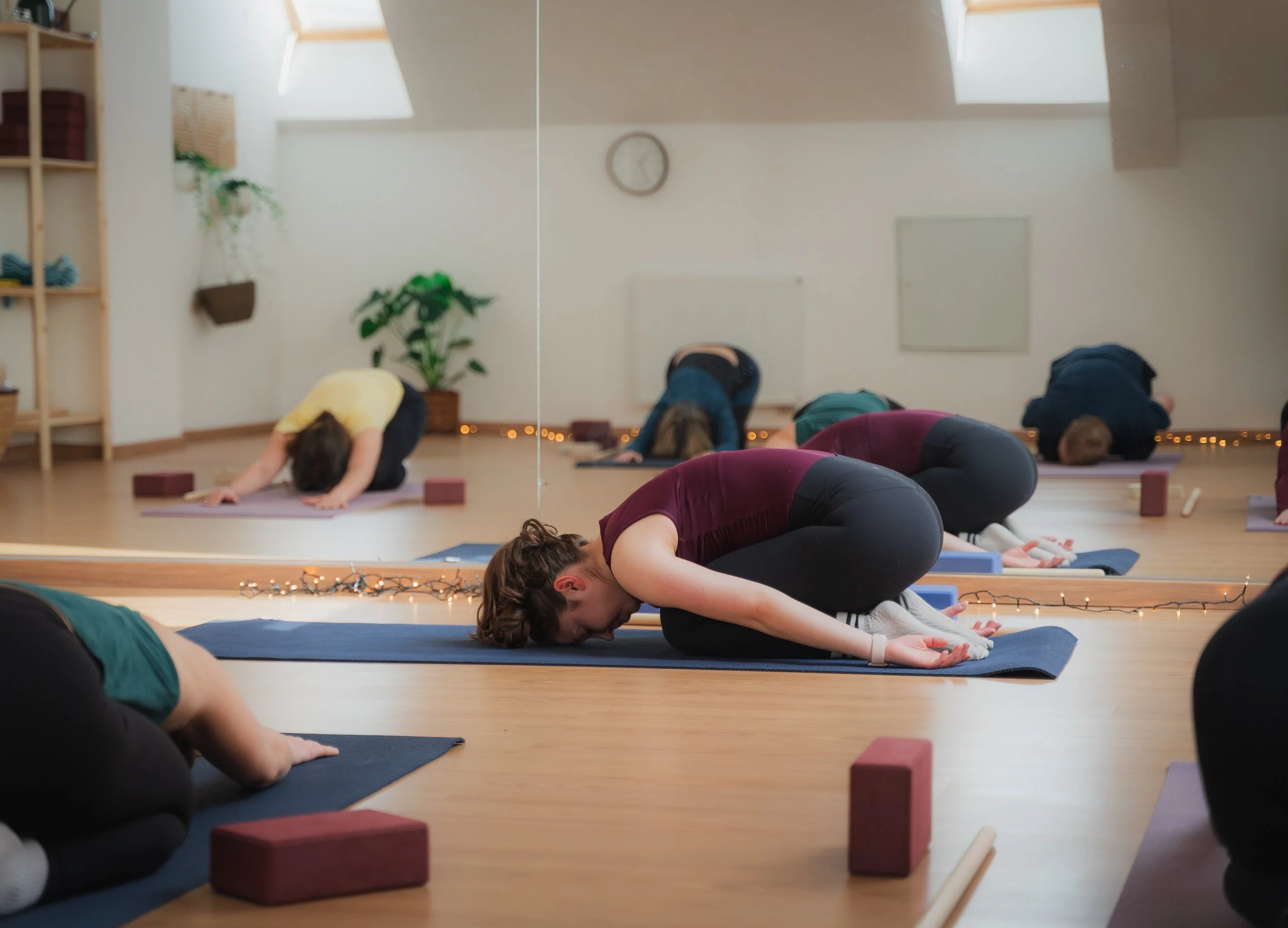 People participating in a yoga class, in child’s pose on yoga mats, with a mirror reflecting the room, wood shelving with baskets, plants, a wall clock, and fairy lights.