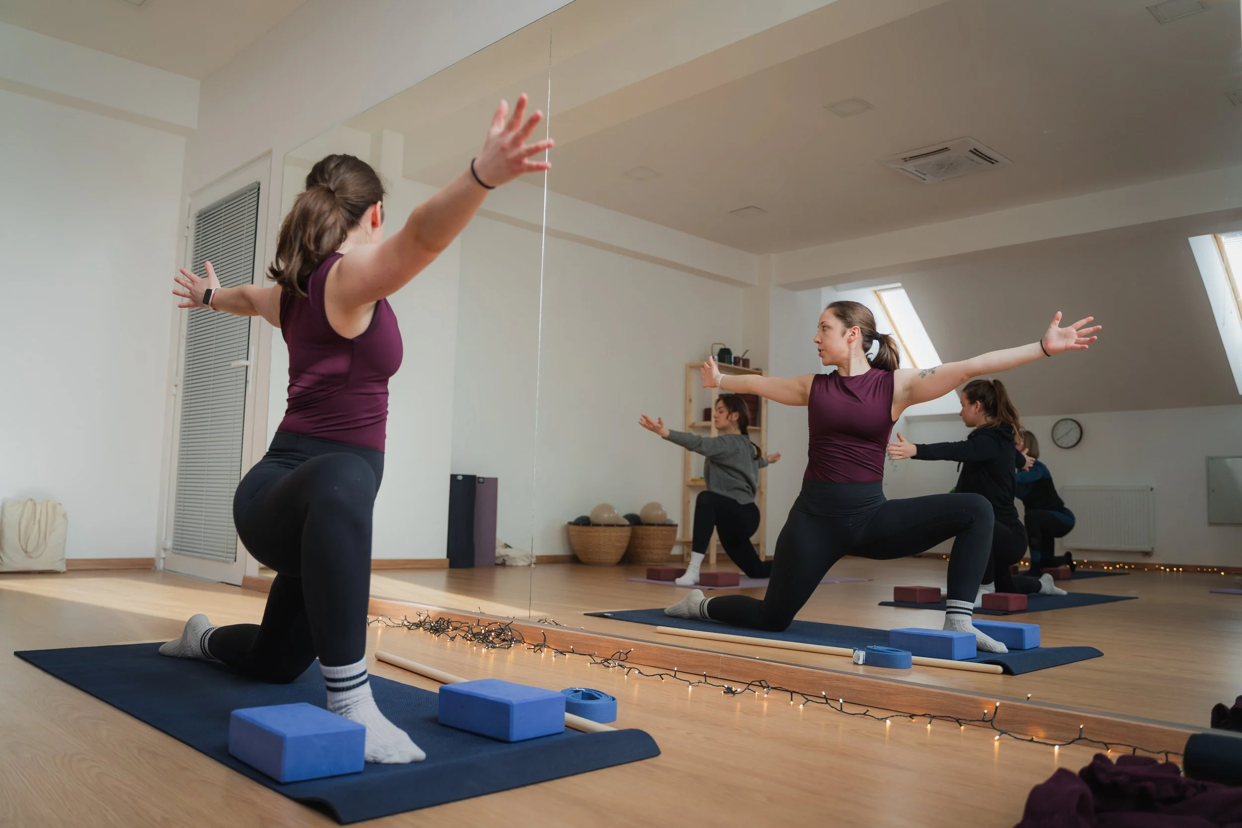 Yoga class with women doing lunges in front of a mirror in a spacious, well-lit studio.