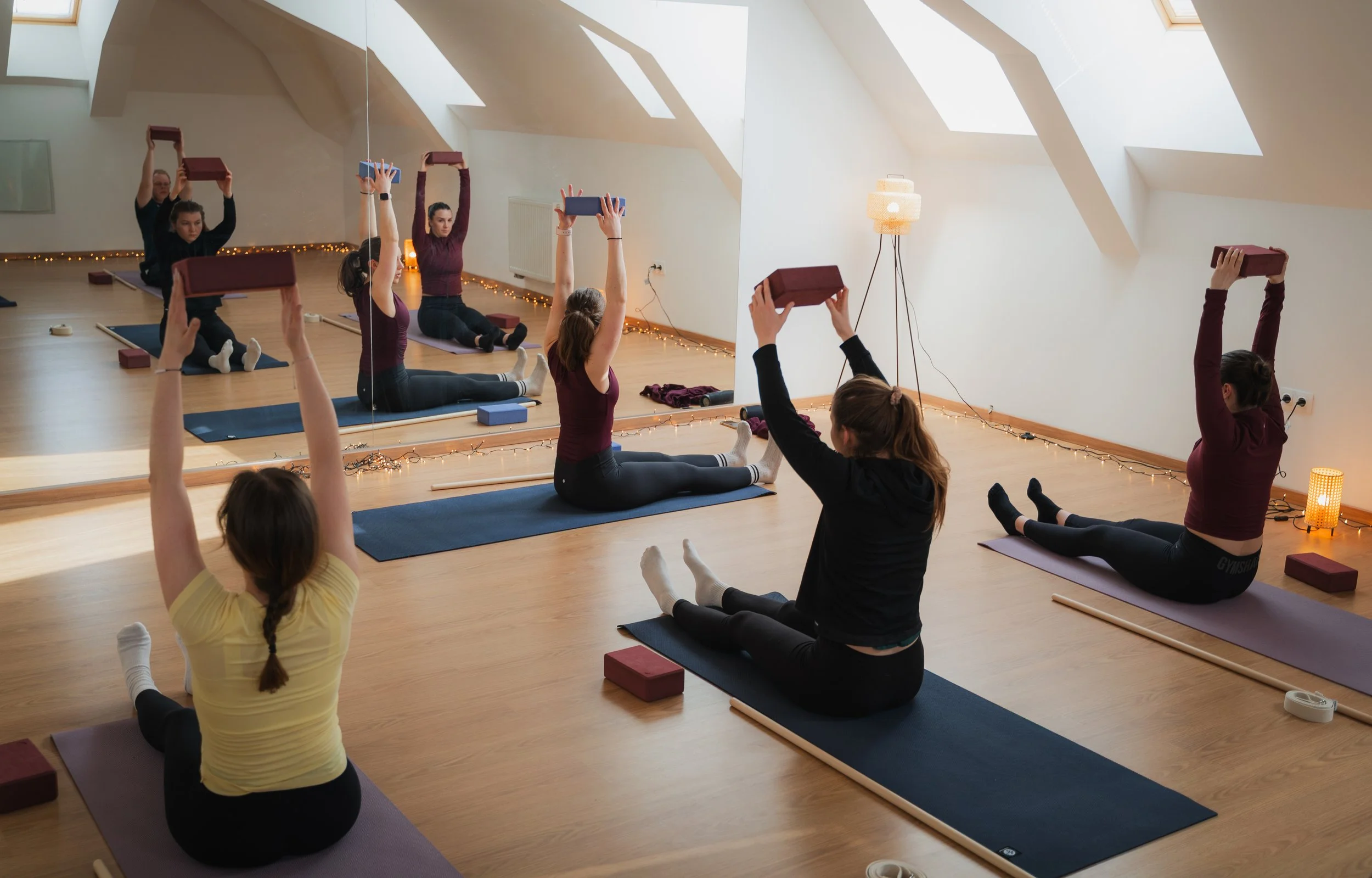 A group of people practicing yoga in a bright, airy studio with wooden floors and mirrored wall, using yoga blocks and mats, with some participants holding weights overhead.