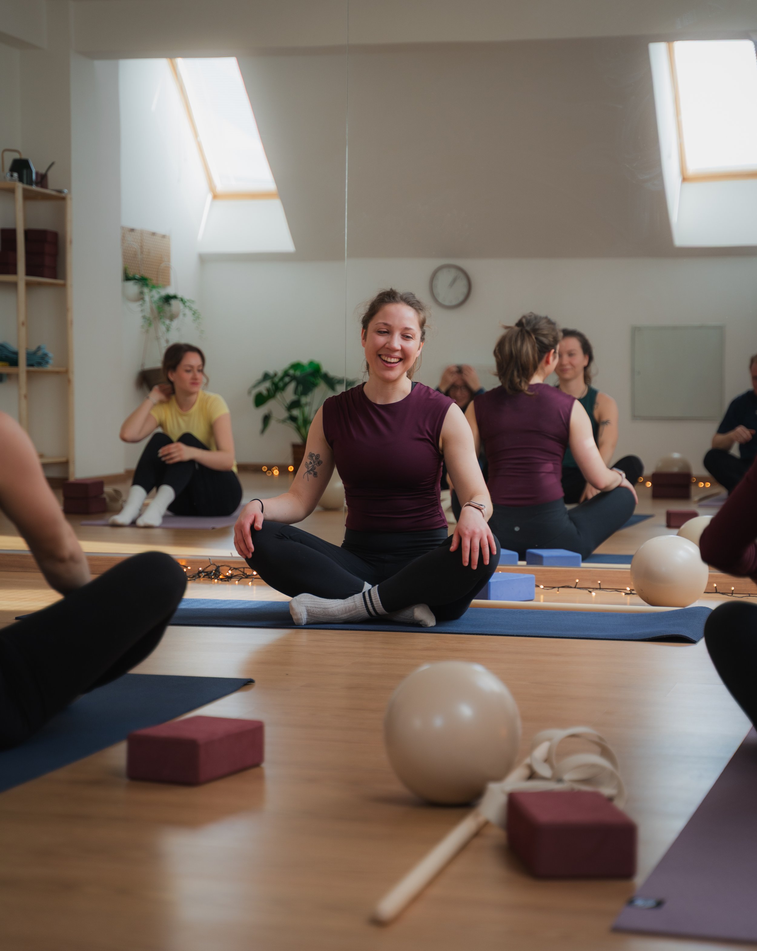 A group of women participating in a yoga class in a studio with natural light, sitting on mats with yoga props around, and one woman smiling at the camera.