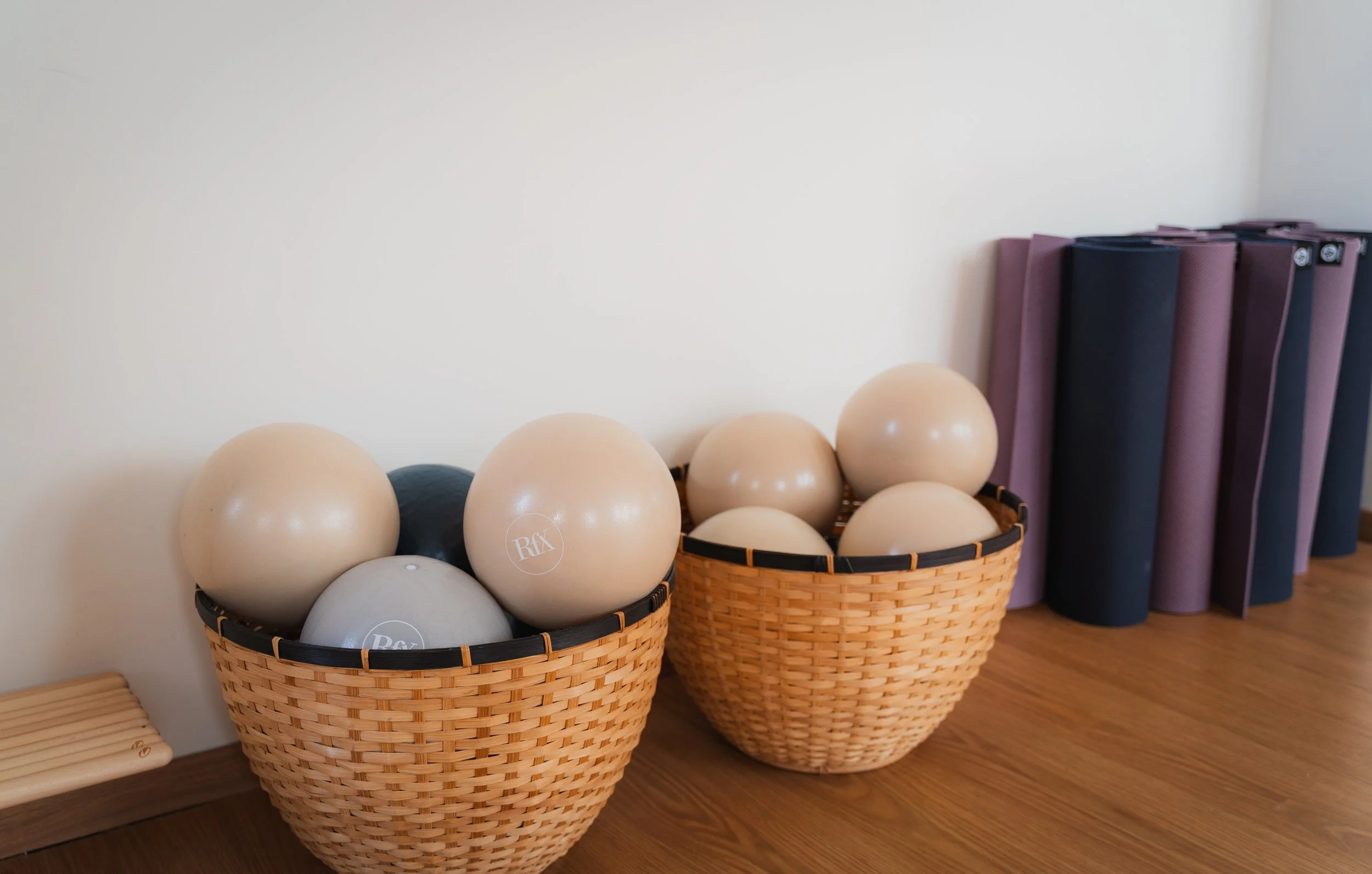 Two wicker baskets filled with exercise balls of various sizes and colors, placed on a wooden floor next to stacked yoga mats.