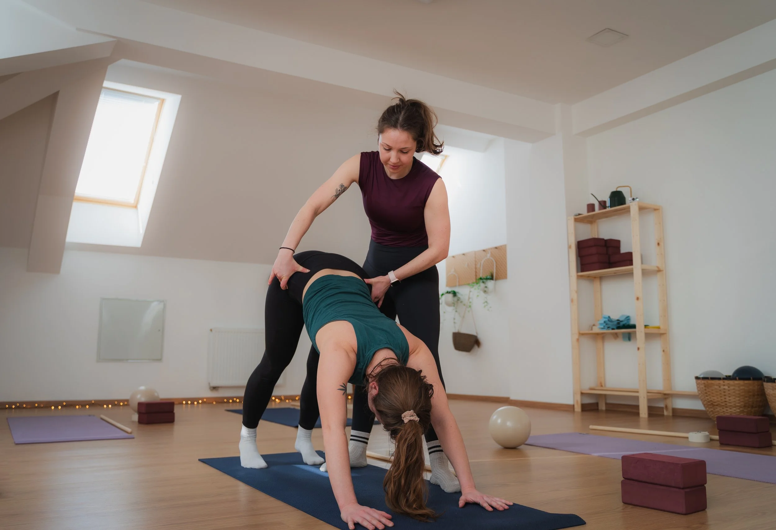 A yoga instructor assisting a woman in a downward dog pose during a yoga class in a bright studio with wooden floor and minimal decor.