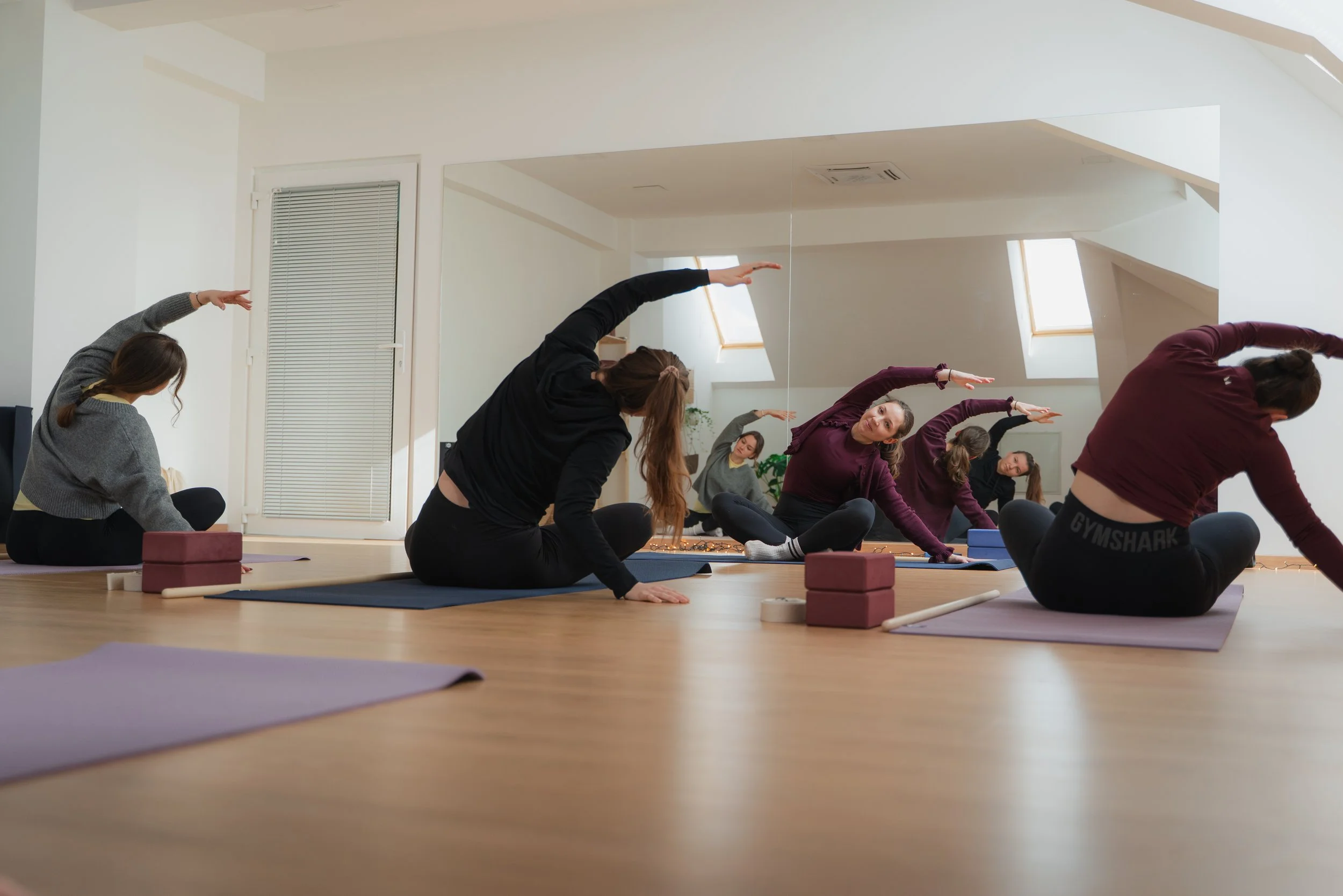 Group of women doing yoga stretches on mats in a bright studio with skylights.