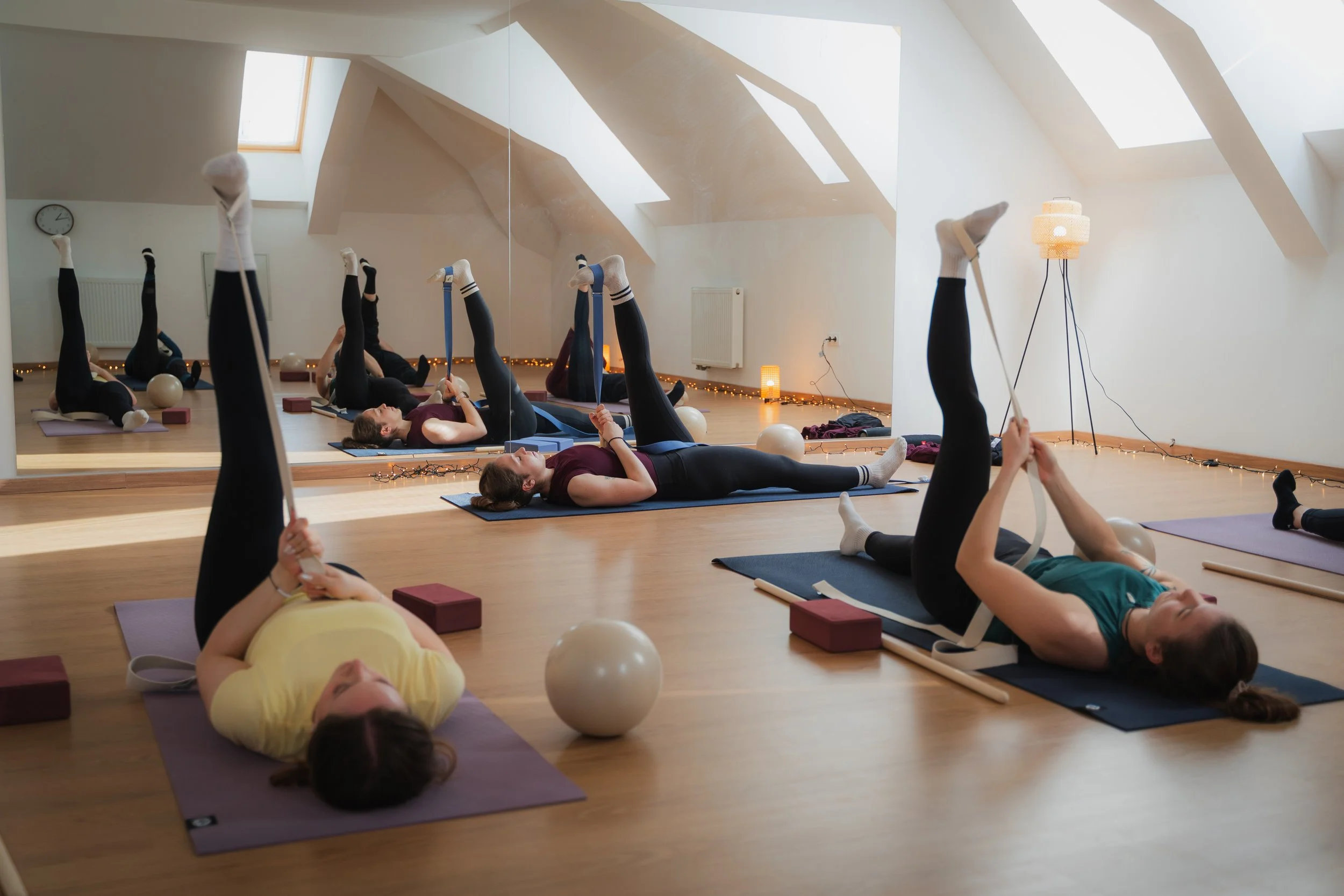 Group of women participating in a Pilates class in a studio with wooden floors, mirrored wall, and skylight windows, lying on yoga mats with resistance bands while engaging in stretching exercises.