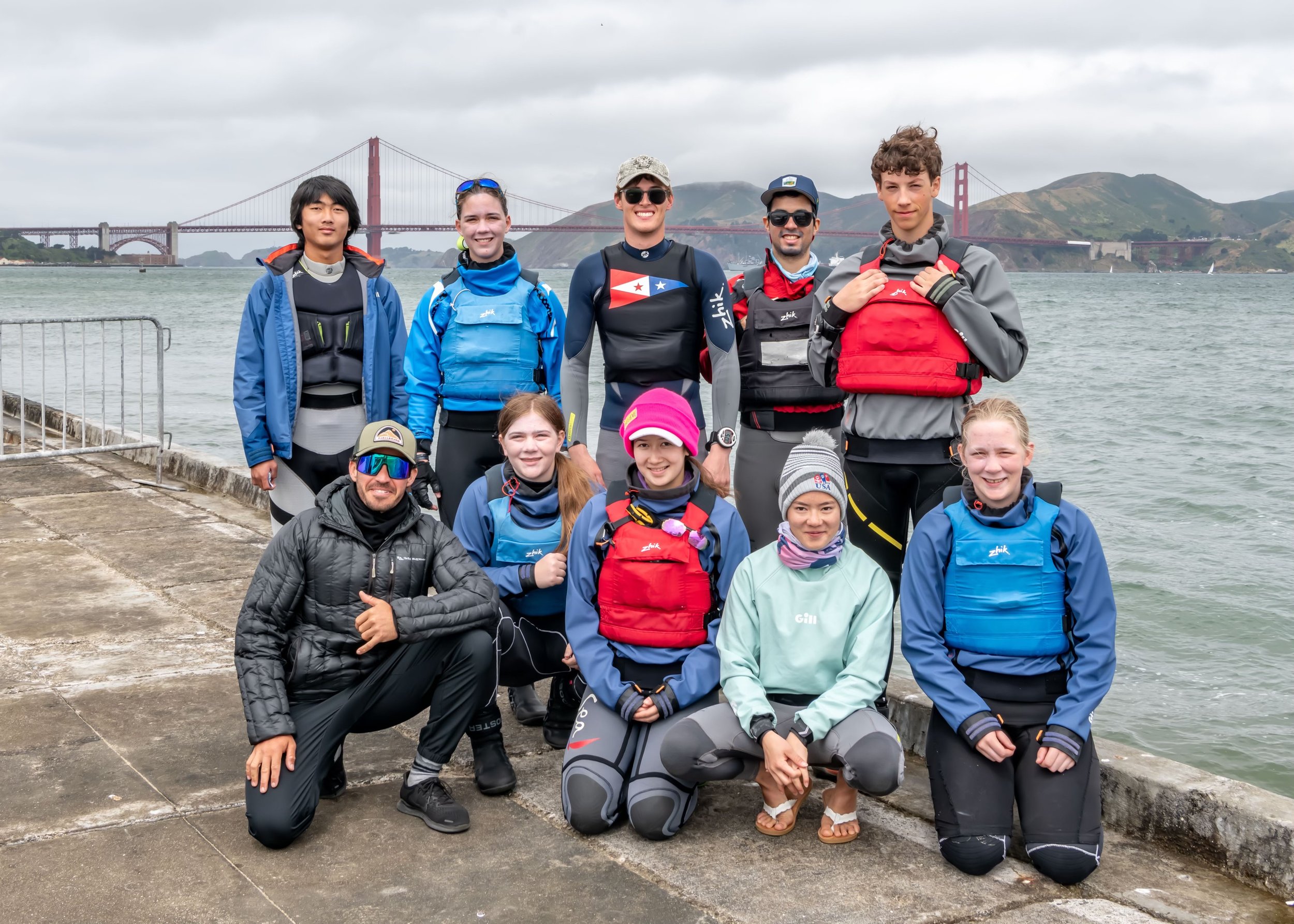 Group of eleven young people in waterproof jackets and backpacks, standing on a concrete pier by the water with the Golden Gate Bridge in the background on a cloudy day.