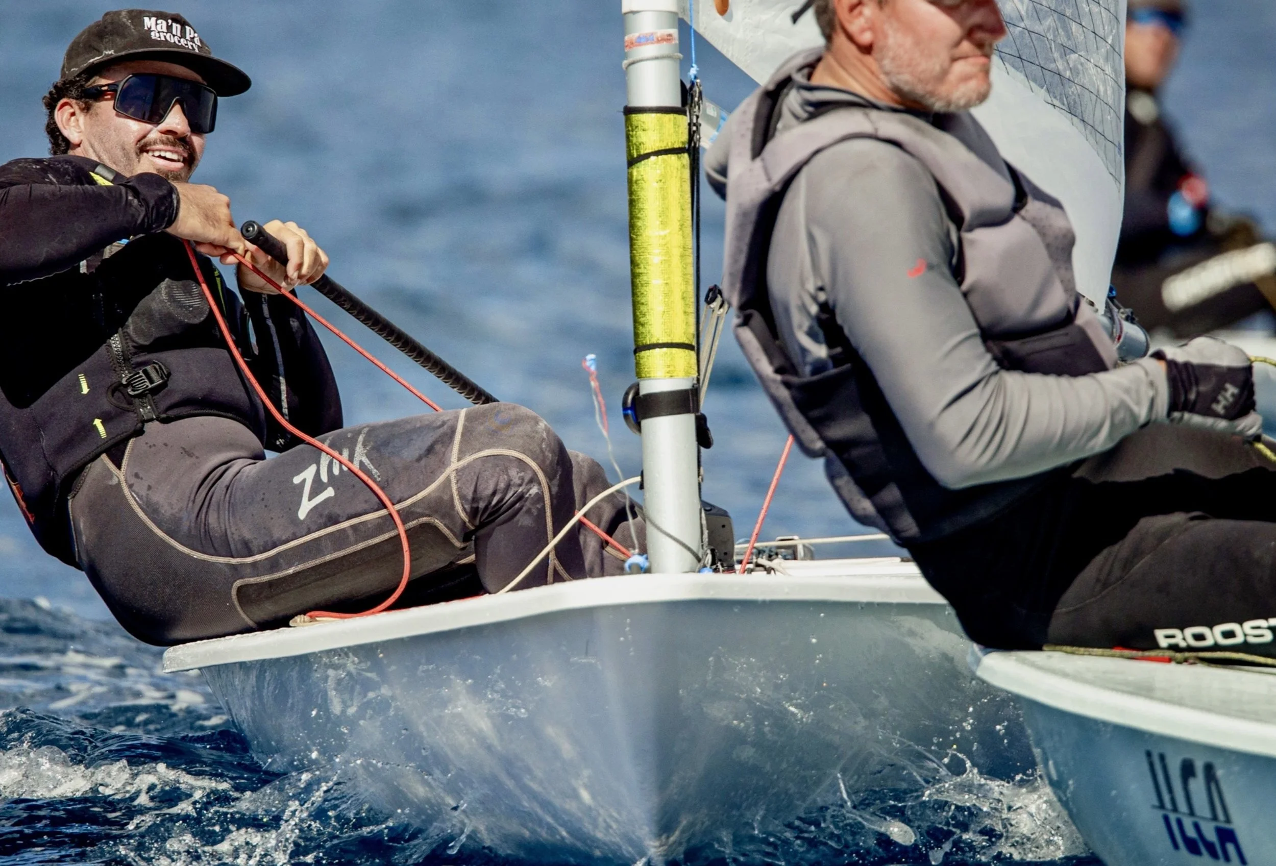 Two men sailing on a small boat, wearing sportswear and sunglasses, with one man smiling and holding a rope, on a water body.