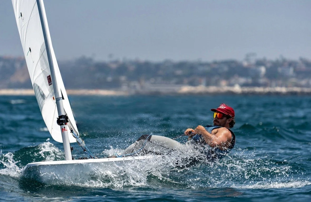 A man sailing a small sailboat on the water, wearing sunglasses and a red cap.