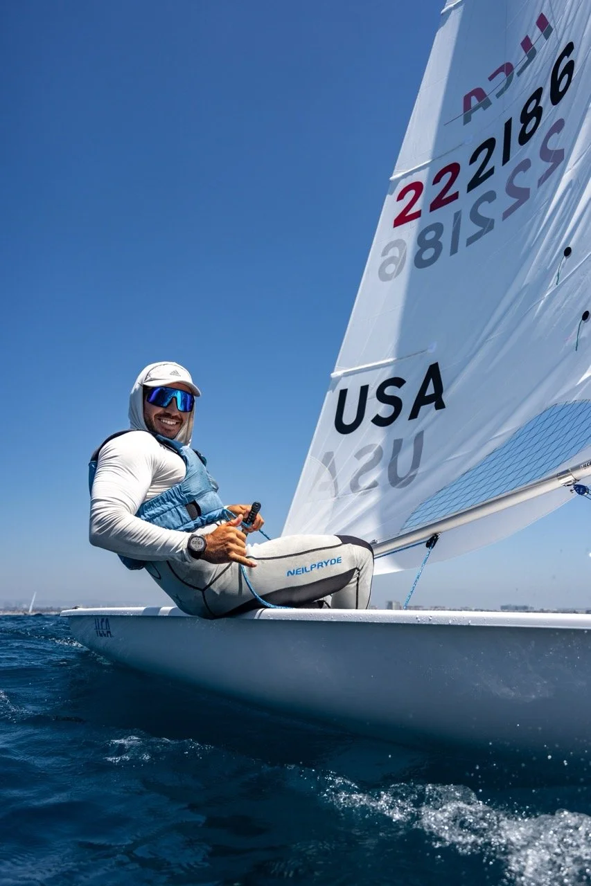 A smiling sailor in sunglasses and a hoodie sailing a small sailboat with an American flag sail, on a clear day with blue sky and water.