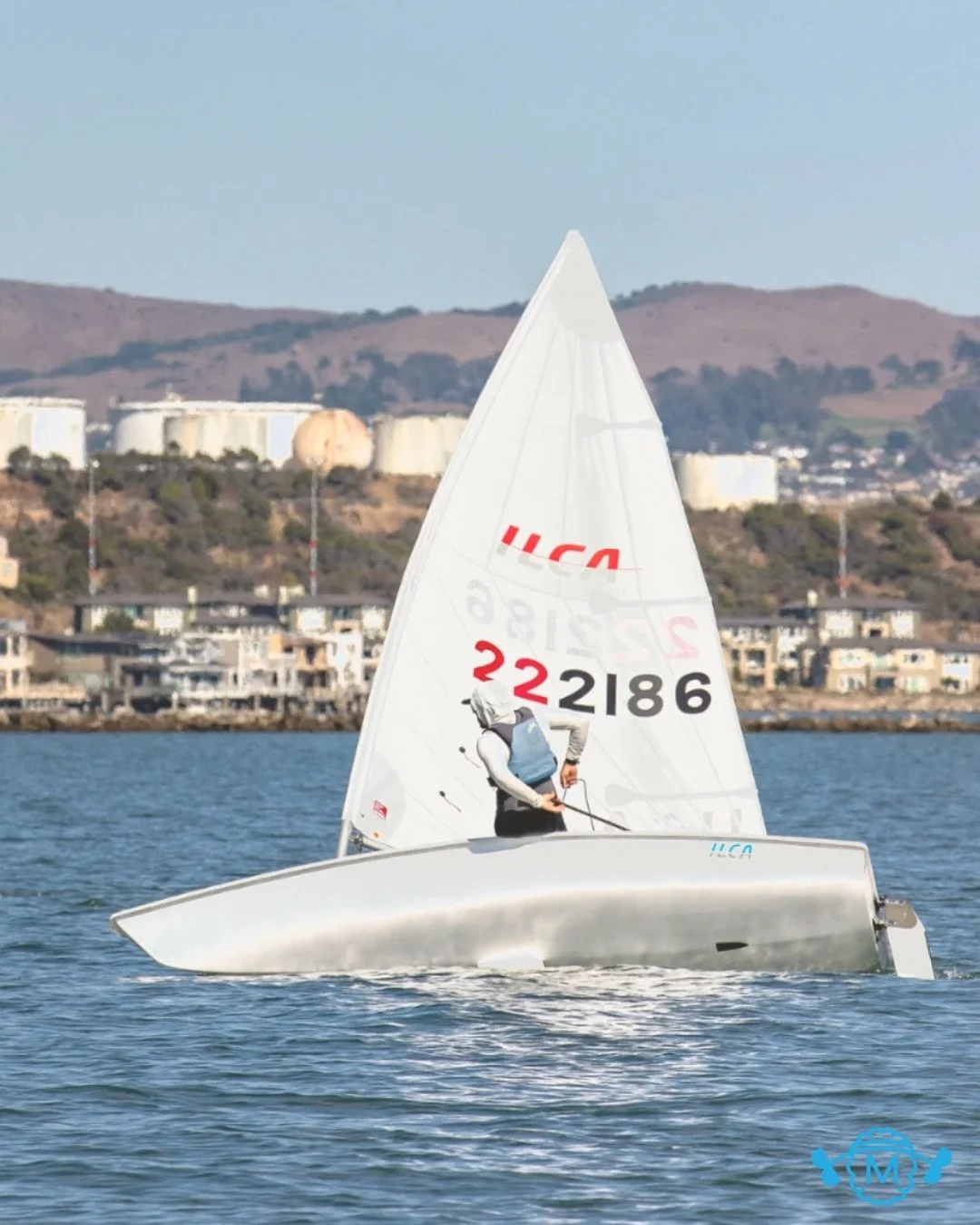 A person sailing a small white sailboat on calm water with a shoreline of houses and industrial tanks in the background.