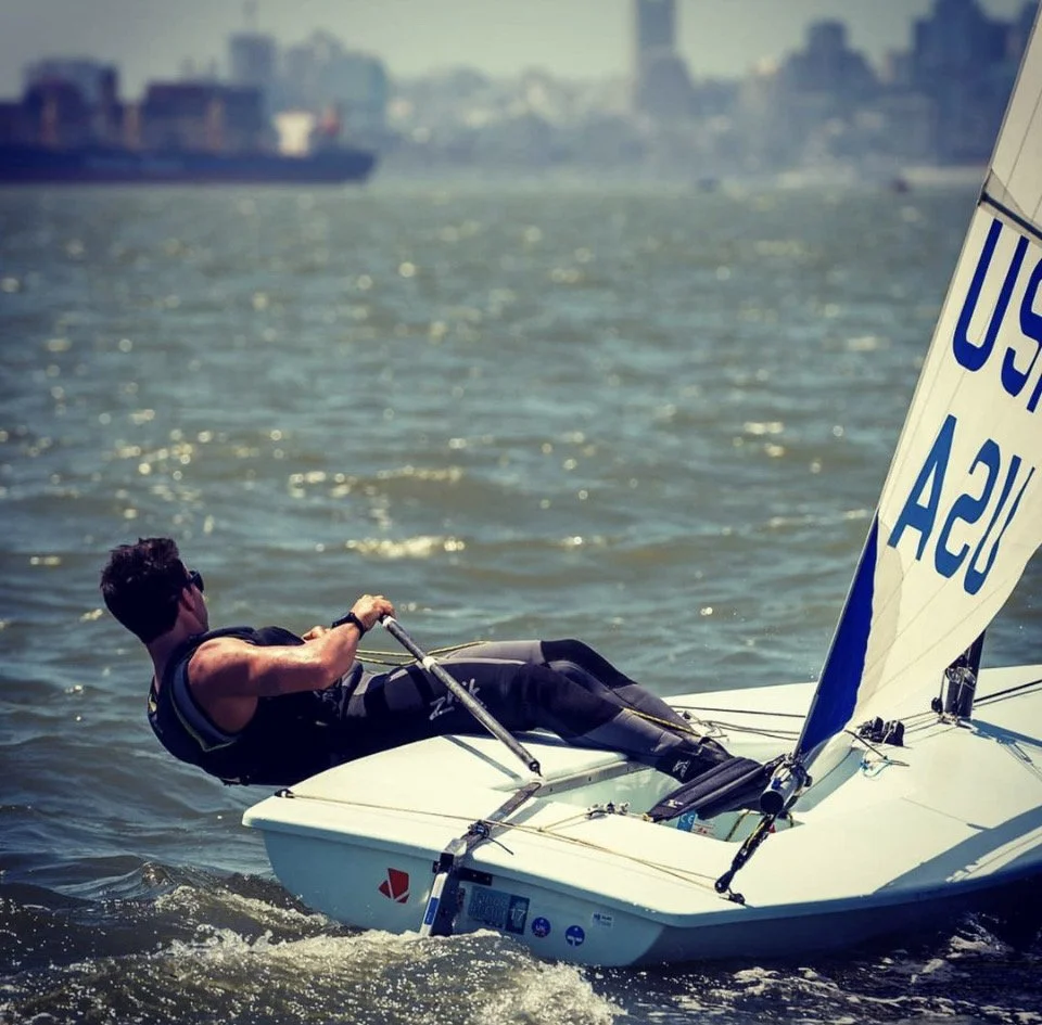 A man sailing a small US Sailing dinghy on a body of water with a city skyline in the background.