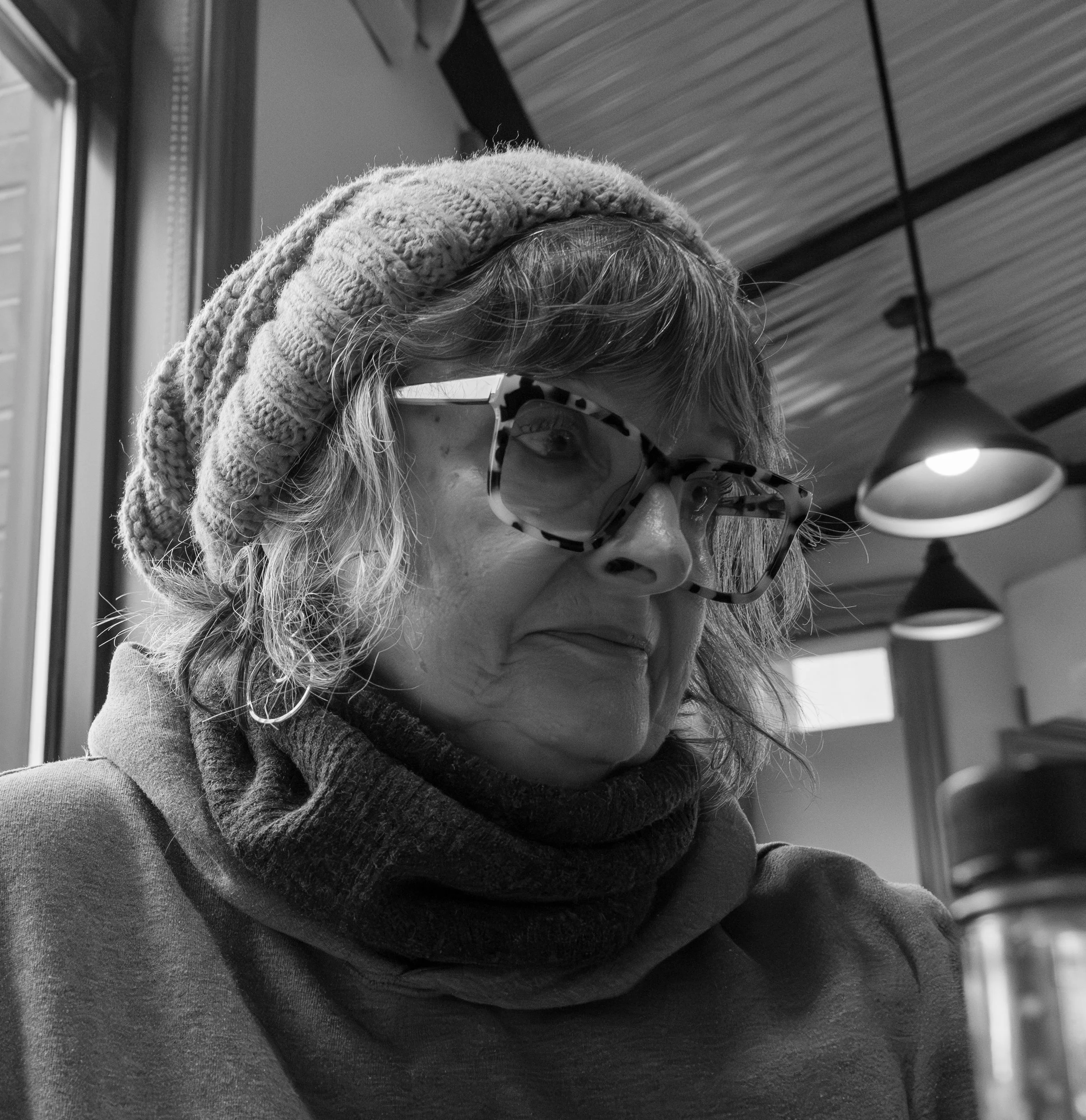 A woman with curly hair wearing a knit cap, large patterned glasses, a scarf, and a hooded sweatshirt, sitting indoors near a window with pendant lights overhead.