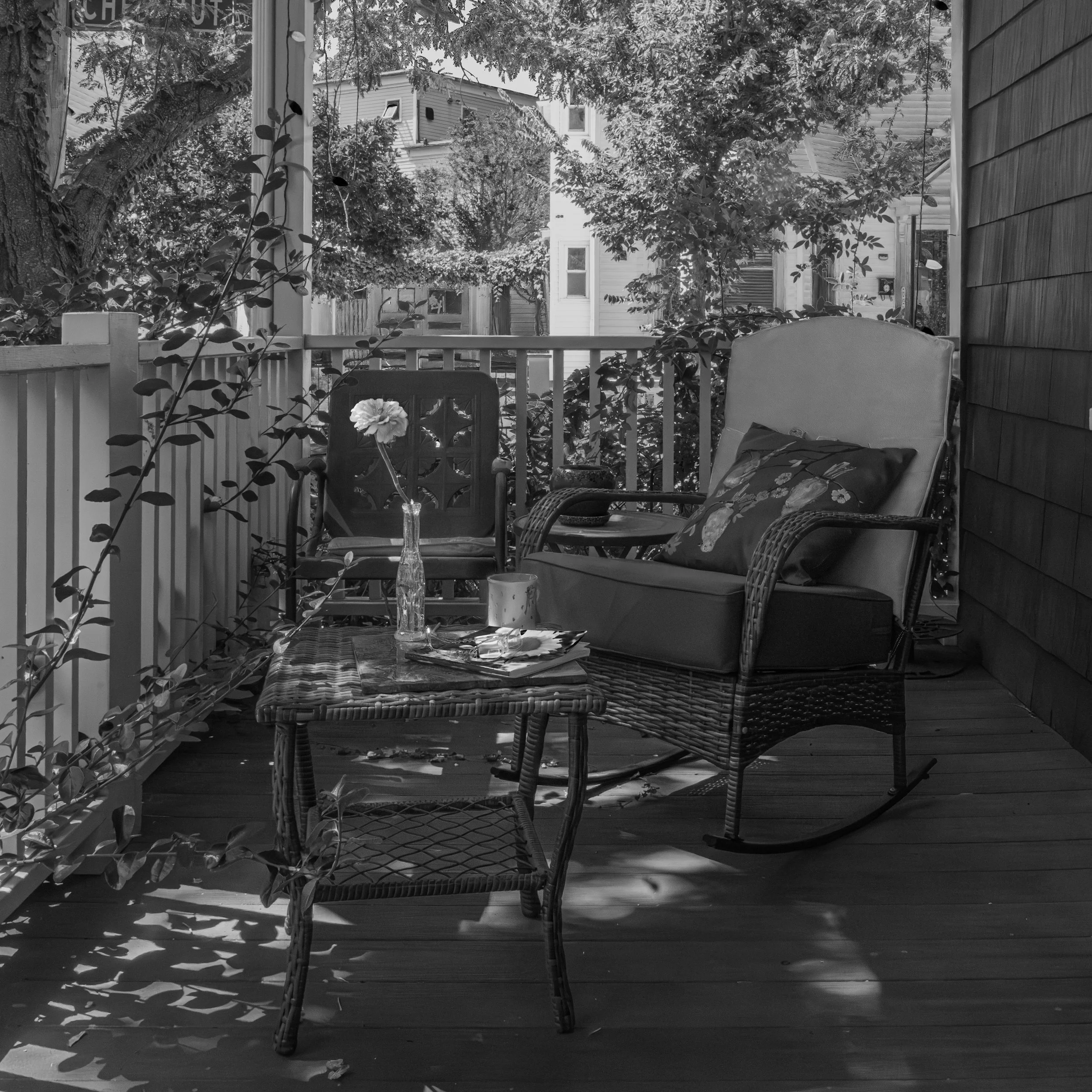 A black and white photo of a cozy outdoor patio with wicker furniture, including a rocking chair and a flowered cushion, a small table with a tray, a vase with a flower, and a decorative cup, surrounded by trees and neighboring houses.