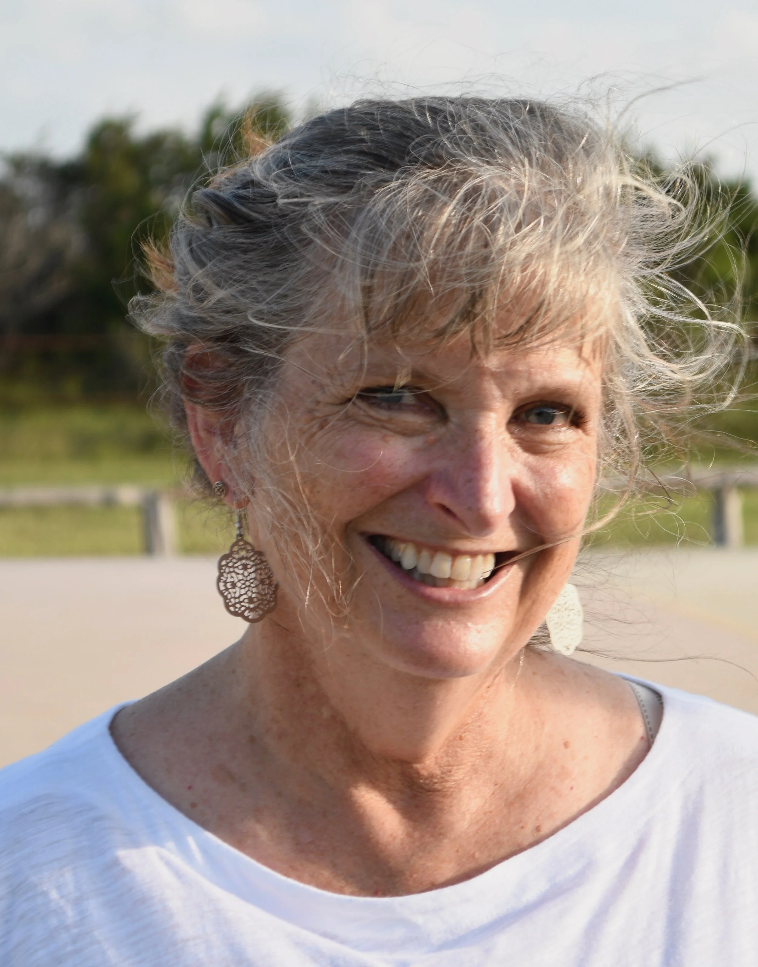 A joyful middle-aged woman with short gray hair, wearing earrings and a white top, smiling outdoors with a blurred natural background.
