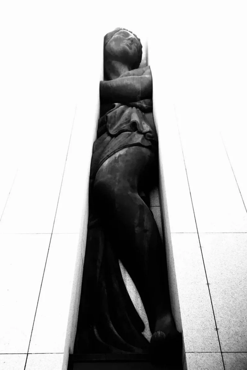 A tall, narrow sculpture of a human face and upper body, with features carved in a dark material, visible within a white, slit-like space.