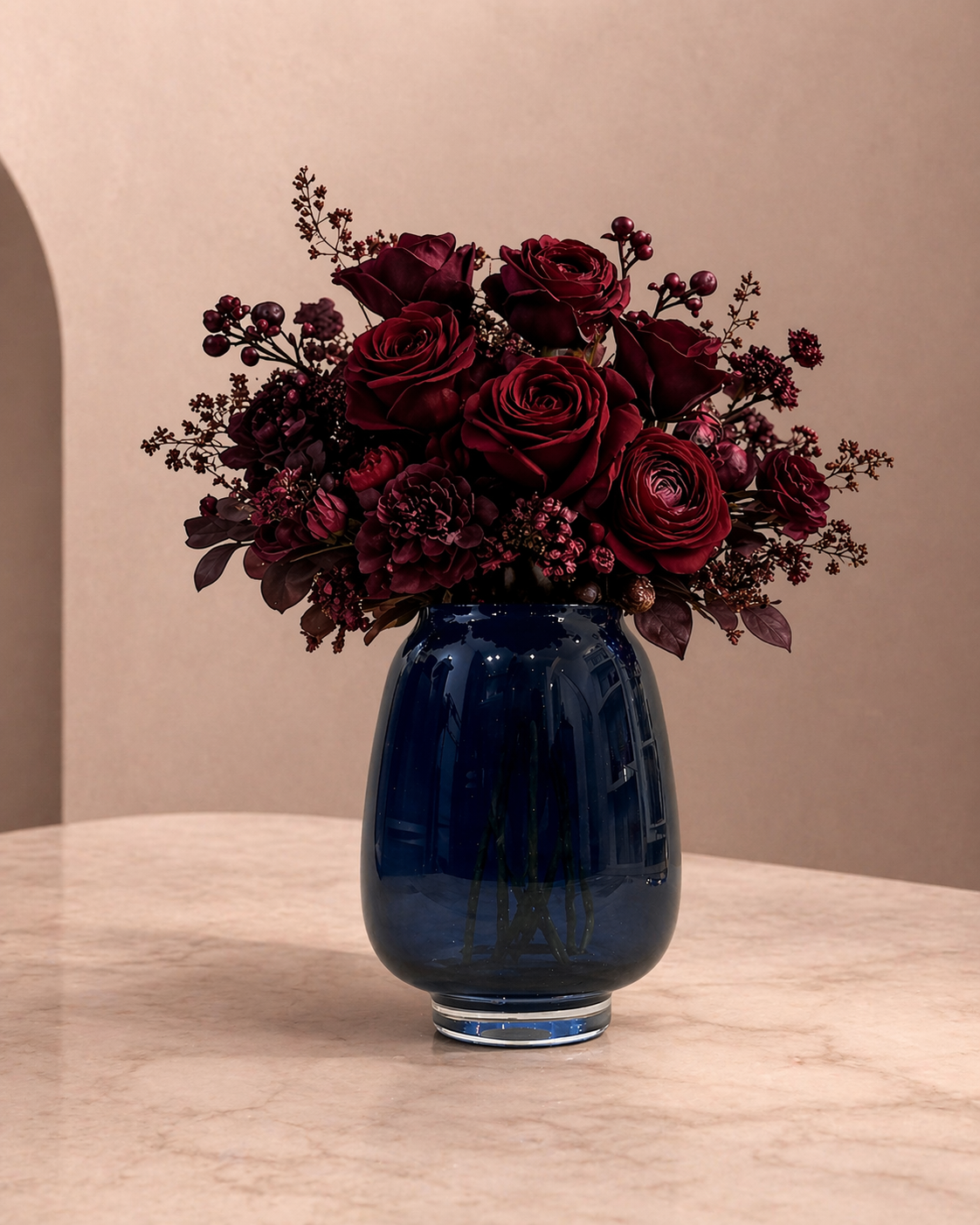 A bouquet of dark red roses and other flowers in a dark blue glass vase on a beige marble surface.