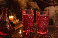 Two glasses filled with red-colored drinks on a table with a candle and a decanter nearby in a dimly lit, cozy setting.