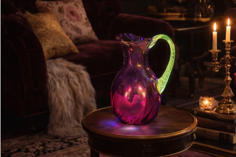 A glowing purple and green glass pitcher on a round wooden table, with candles and a sofa in the background.