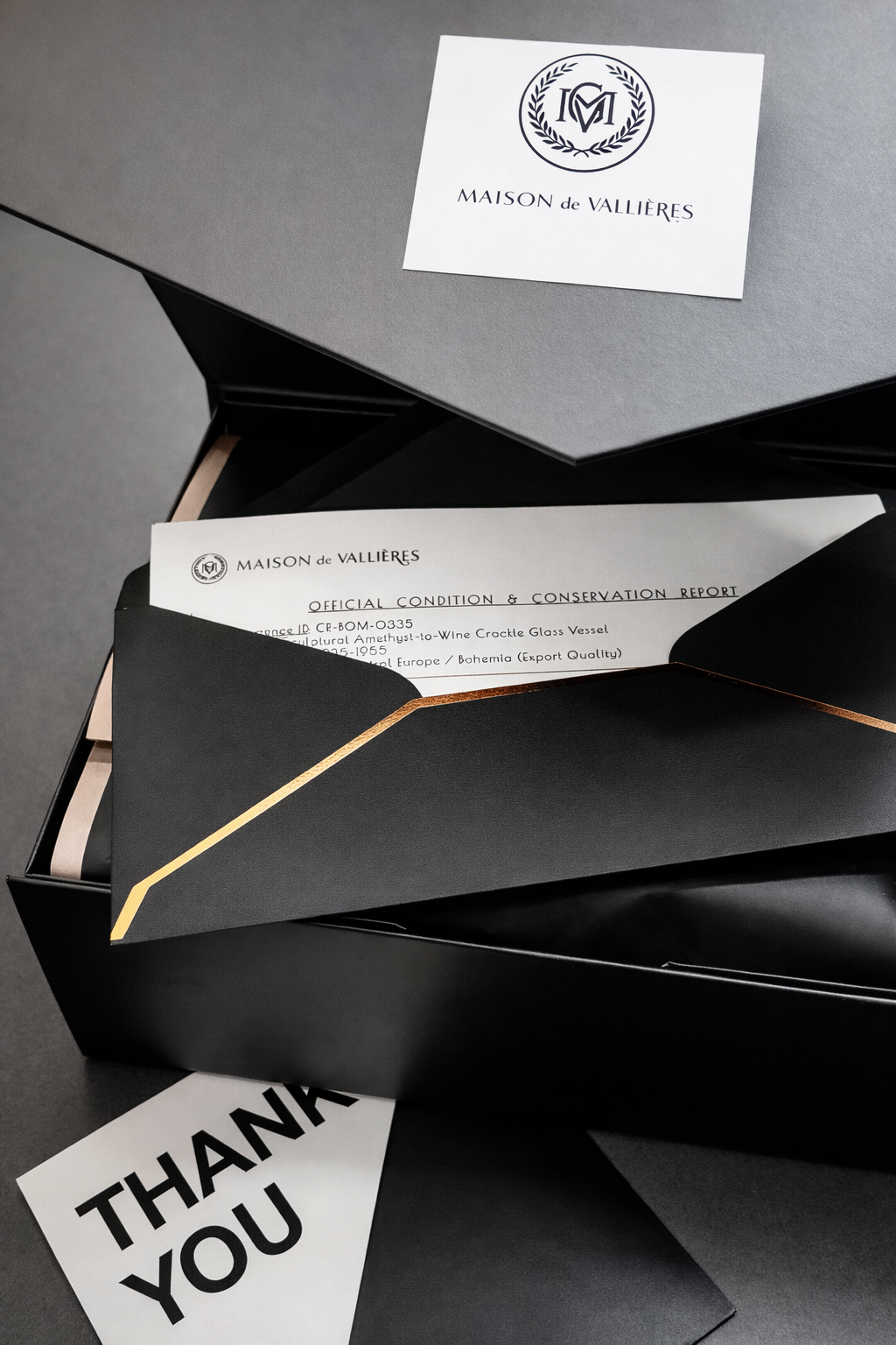 A black gift box with a lid featuring a white and navy logo of Maison de Vallières, with a document titled 'Official Condition & Conservation Report' partially visible inside, a black envelope with a gold stripe, and a white thank you card.