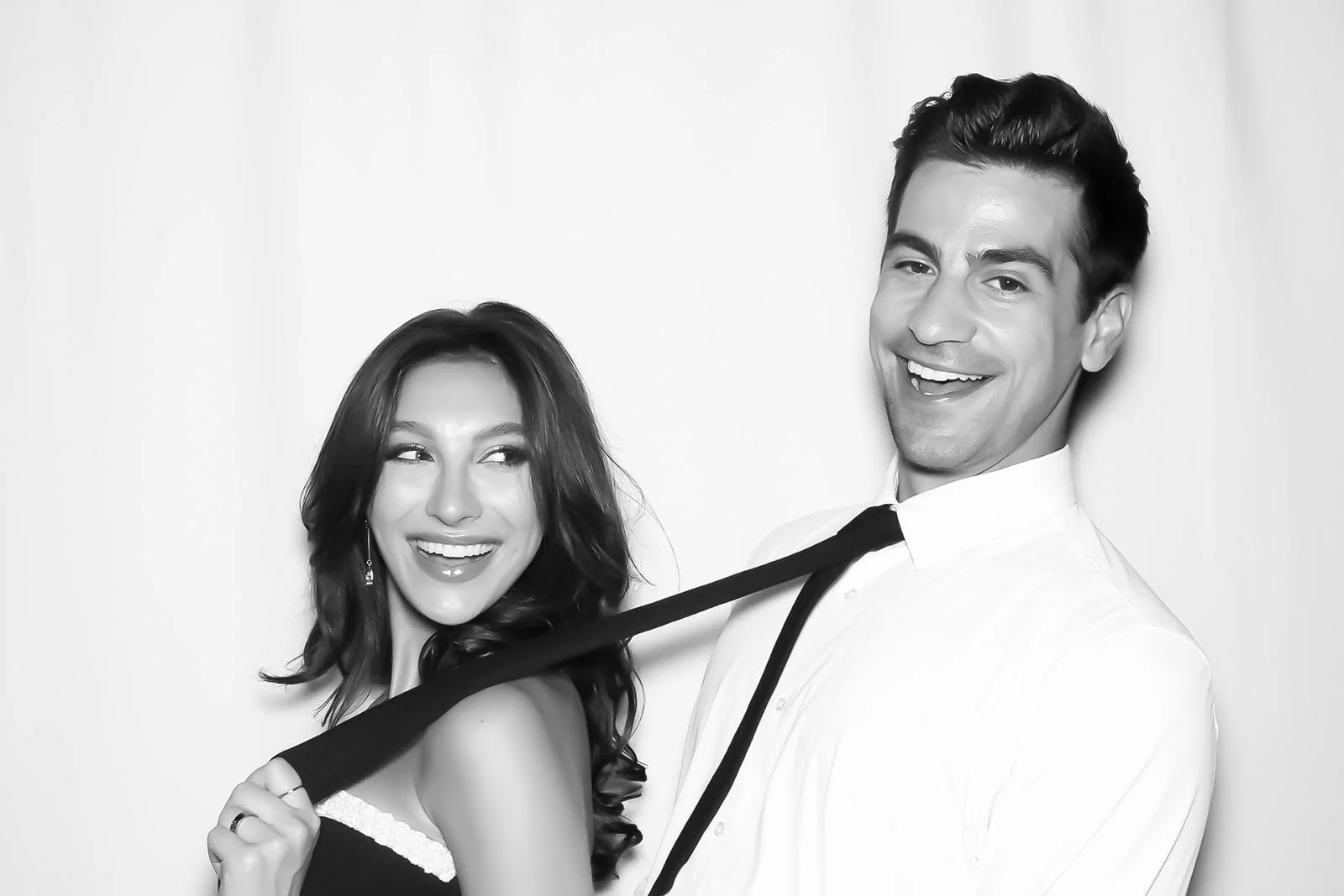 A black and white photo of a smiling man in a white shirt and black tie and a woman in a dark dress with white trim, with the woman holding the man's tie.