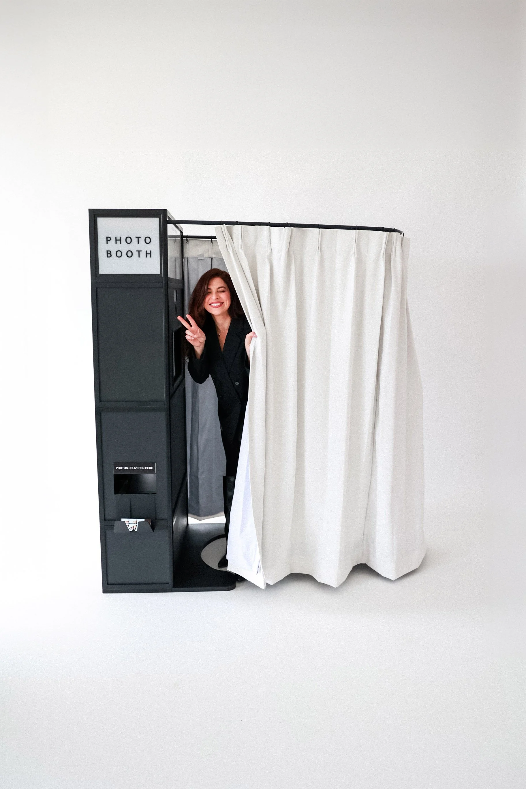 Woman smiling and making a peace sign while peeking out of a photo booth with white curtains, on a plain white background.
