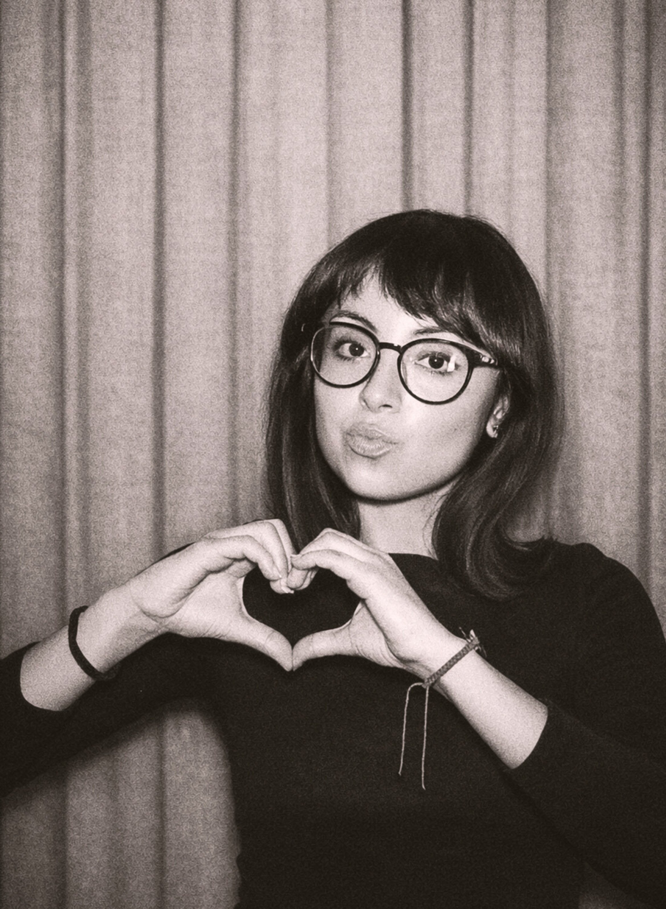 Black and white photo of a woman with glasses and shoulder-length hair making a heart shape with her hands.