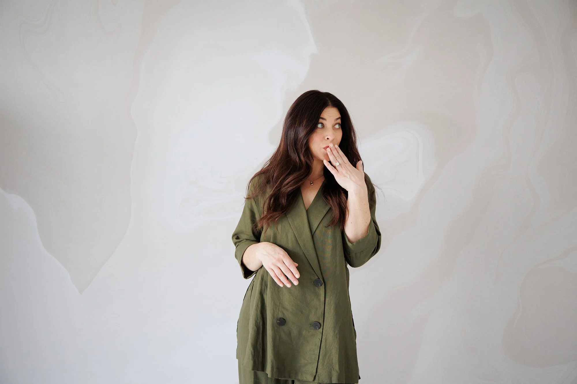 Woman with long brown hair, wearing an olive green blazer, standing against a light patterned wall, with her hand near her mouth, appearing surprised or thoughtful.