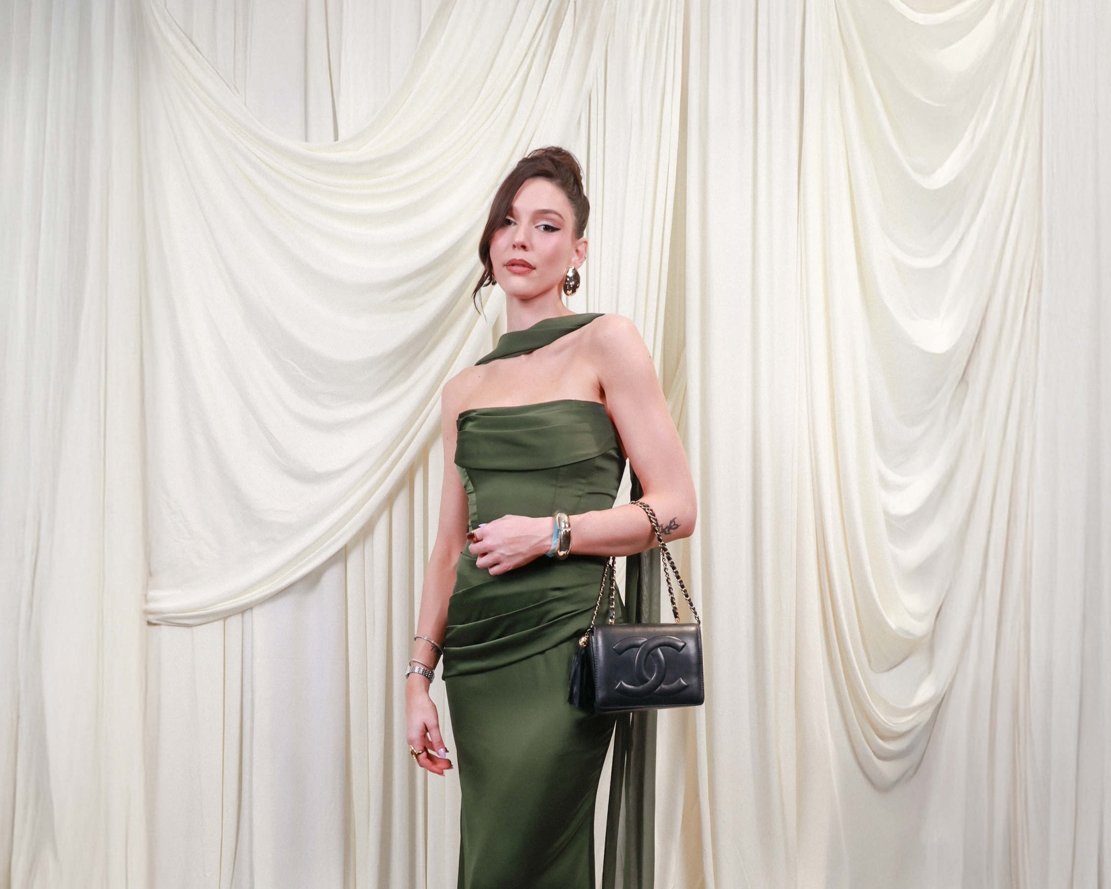 Woman in an elegant green dress holding a chain-strap black Chanel purse standing in front of cream-colored drapery.