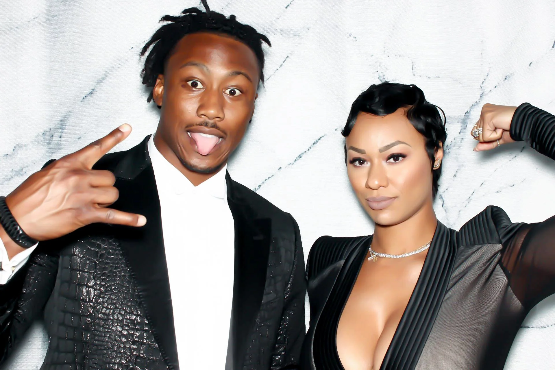 Two people, a man and a woman, posing together in front of a white marble background. The man is wearing a black suit with a white shirt and making a playful face with his tongue out and his right hand making a rock and roll gesture. The woman has short black hair, is dressed in a black outfit with sheer sleeves, and is showing her bicep with her right arm flexed.