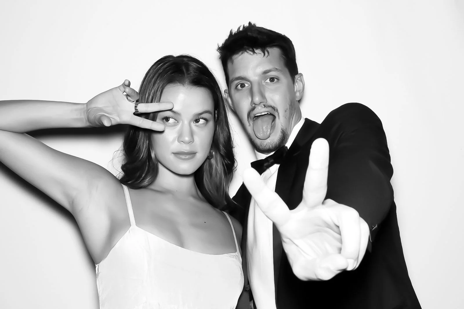 Black and white photo of a young woman and man posing playfully; the woman is making a peace sign near her eye, and the man is sticking his tongue out and holding up a peace sign.