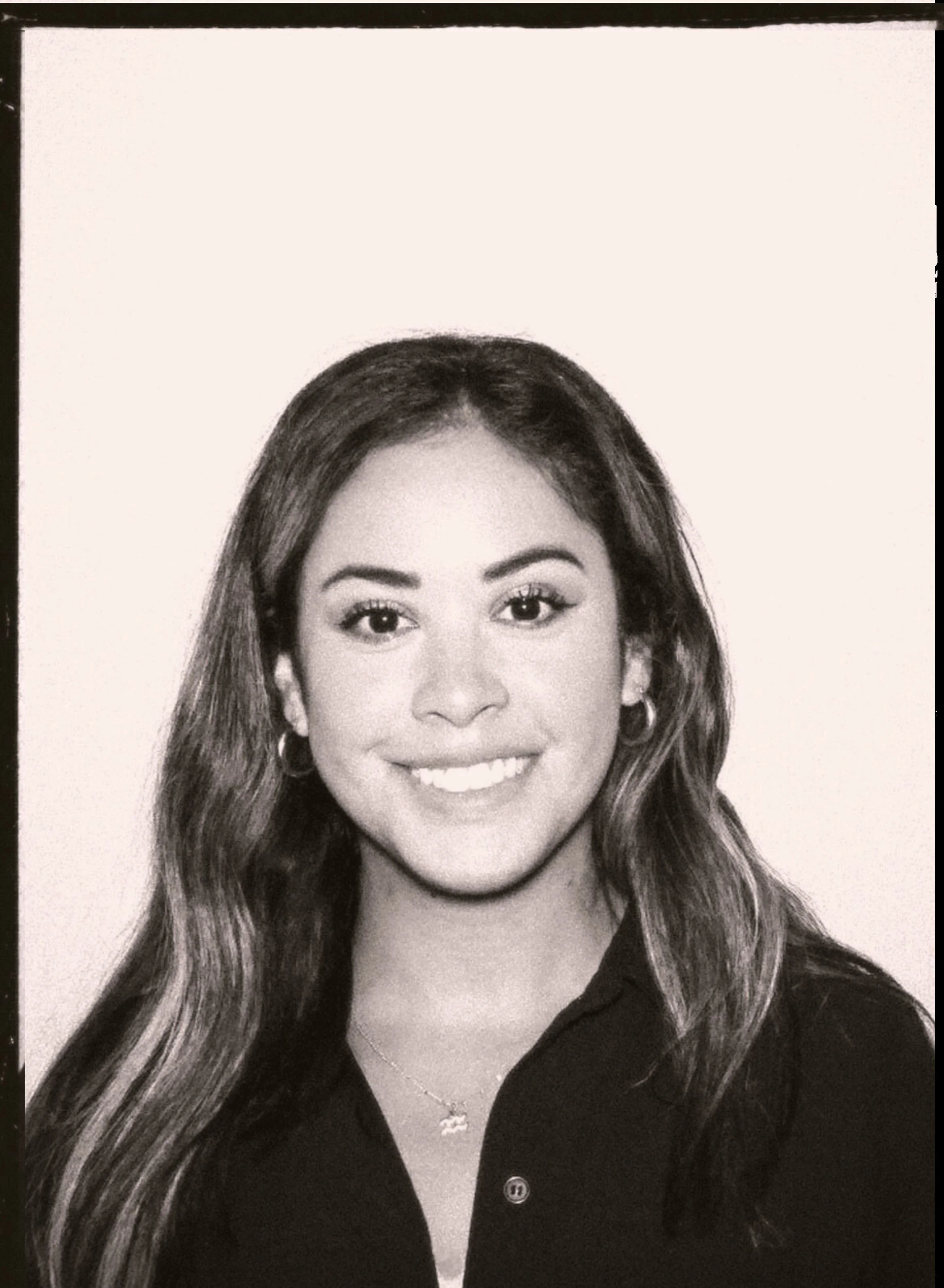Black and white portrait of a young woman with long hair, smiling, wearing hoop earrings, a necklace, and a collared shirt.