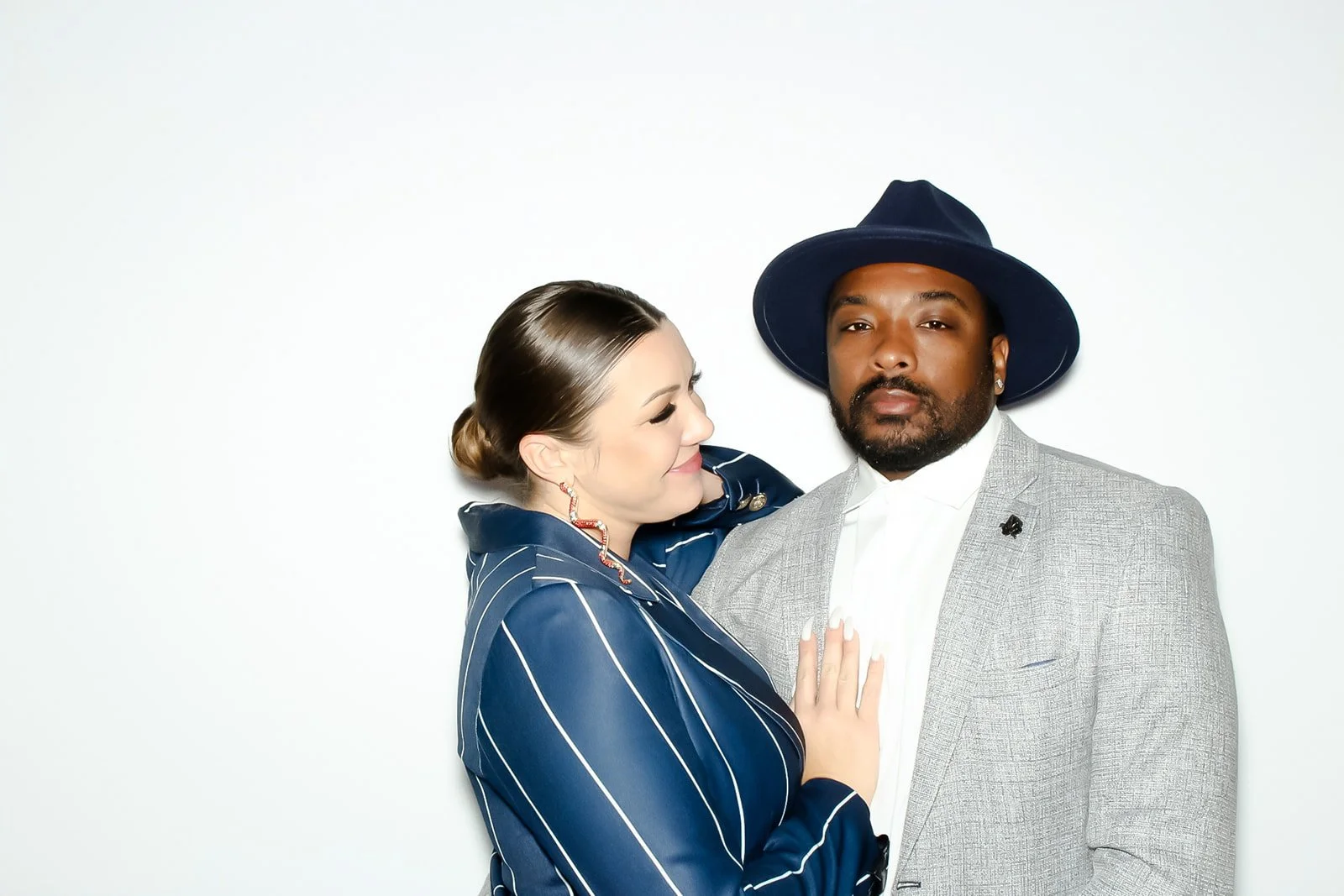 A woman with makeup and earrings standing close to a man wearing a gray blazer, a white shirt, and a large navy blue hat, against a plain white background.