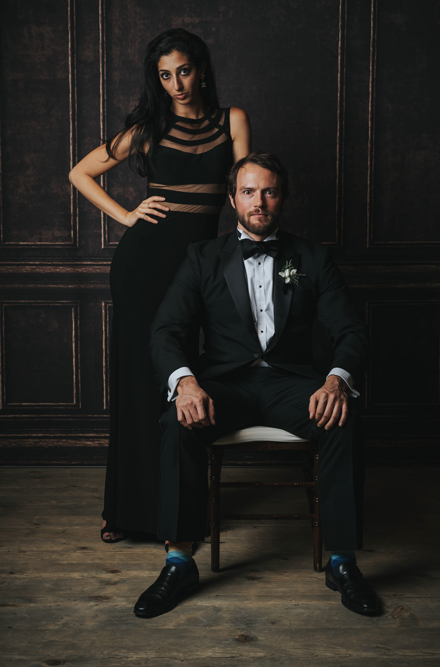A man in a tuxedo sitting on a chair and a woman in a black gown standing behind him with her hand on her hip, all against a dark wooden panel background.