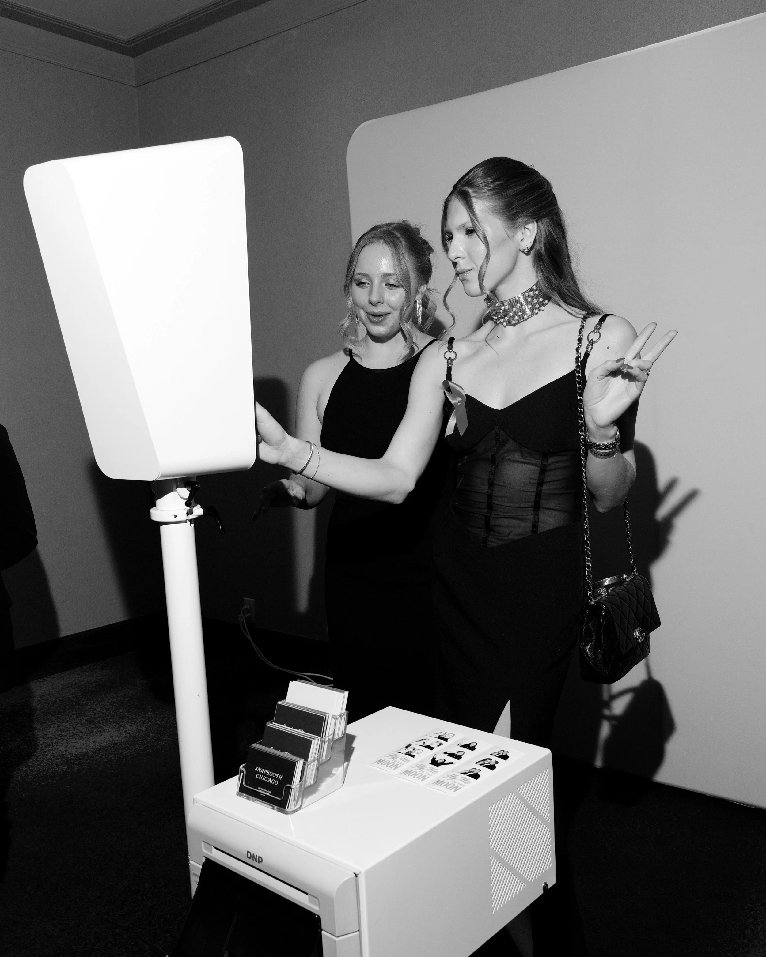 Two women in elegant black dresses taking a selfie at a photo booth, with one woman making a peace sign gesture.