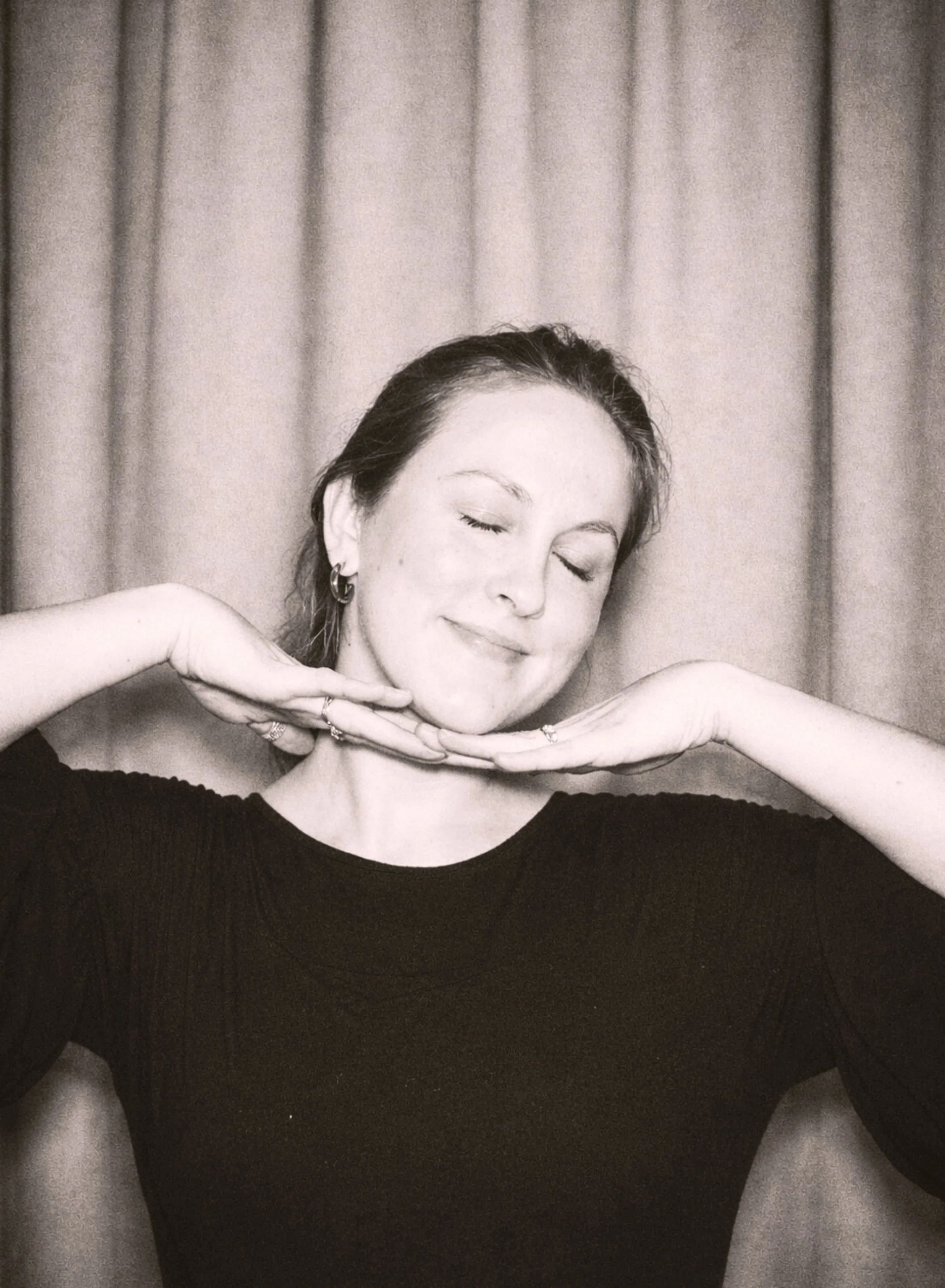 Black and white photo of a woman with closed eyes and a gentle smile, resting her chin on her hands, against a curtain background.