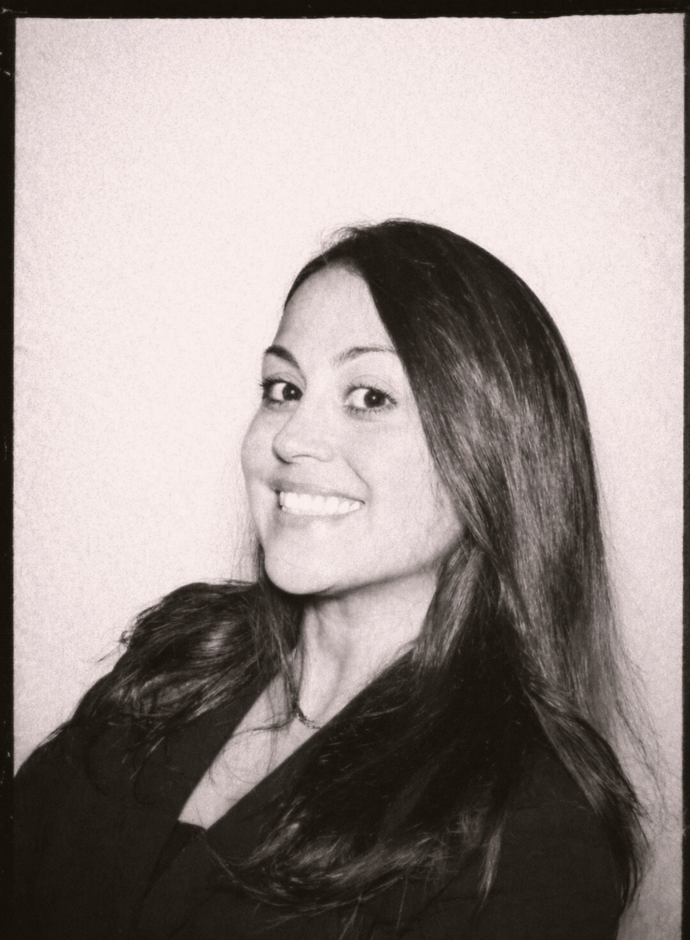 Black and white portrait of a woman with long dark hair, smiling, looking over her shoulder with a plain background.