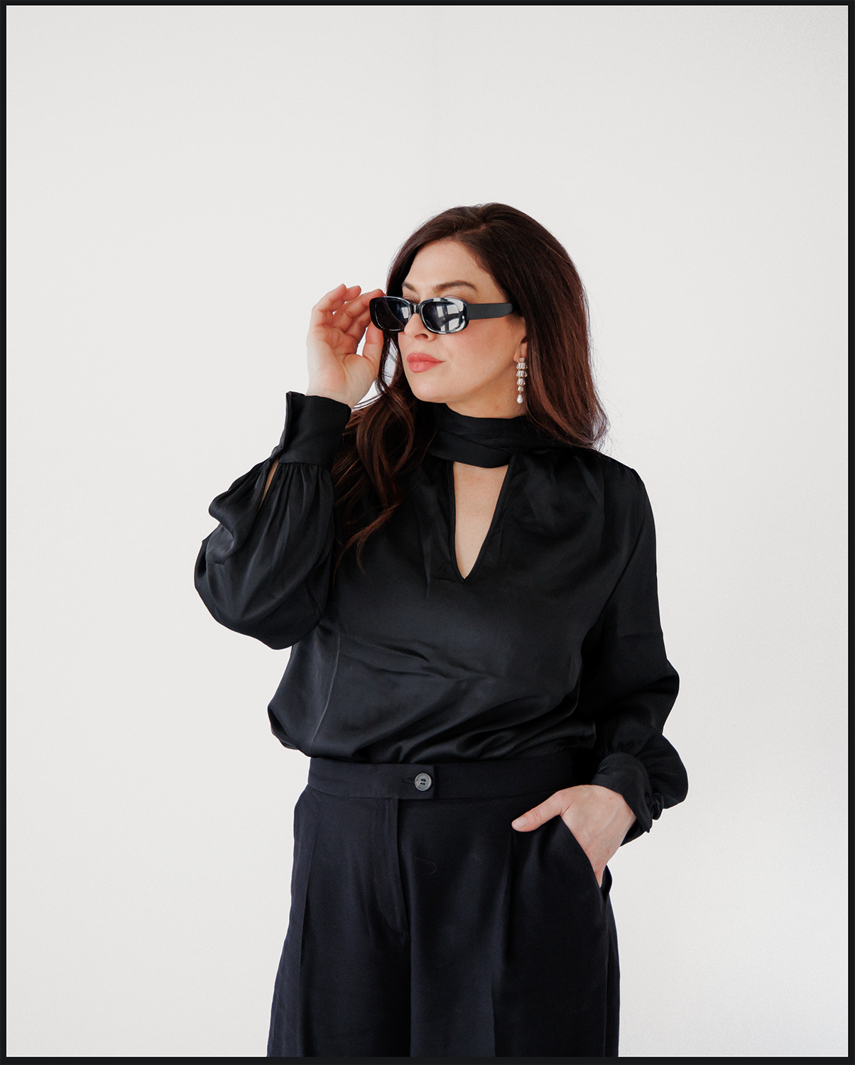 A woman with long dark hair wearing black sunglasses, a black satin blouse, matching black pants, and dangling earrings, standing against a plain white background.