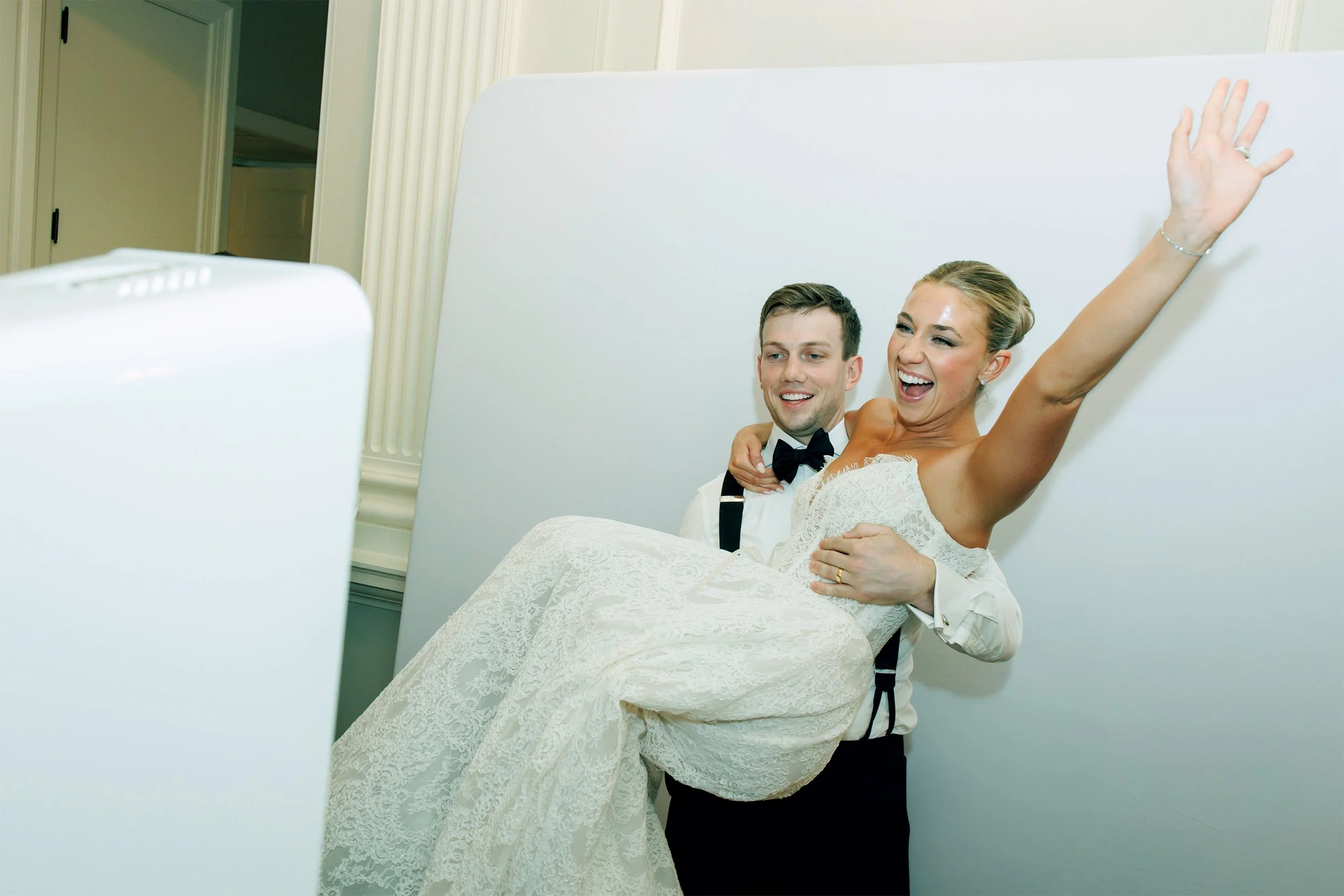 A newlywed couple taking a joyful selfie, the bride is sitting on the groom's lap waving and smiling, dressed in a wedding gown, the groom in a tuxedo with a bow tie.