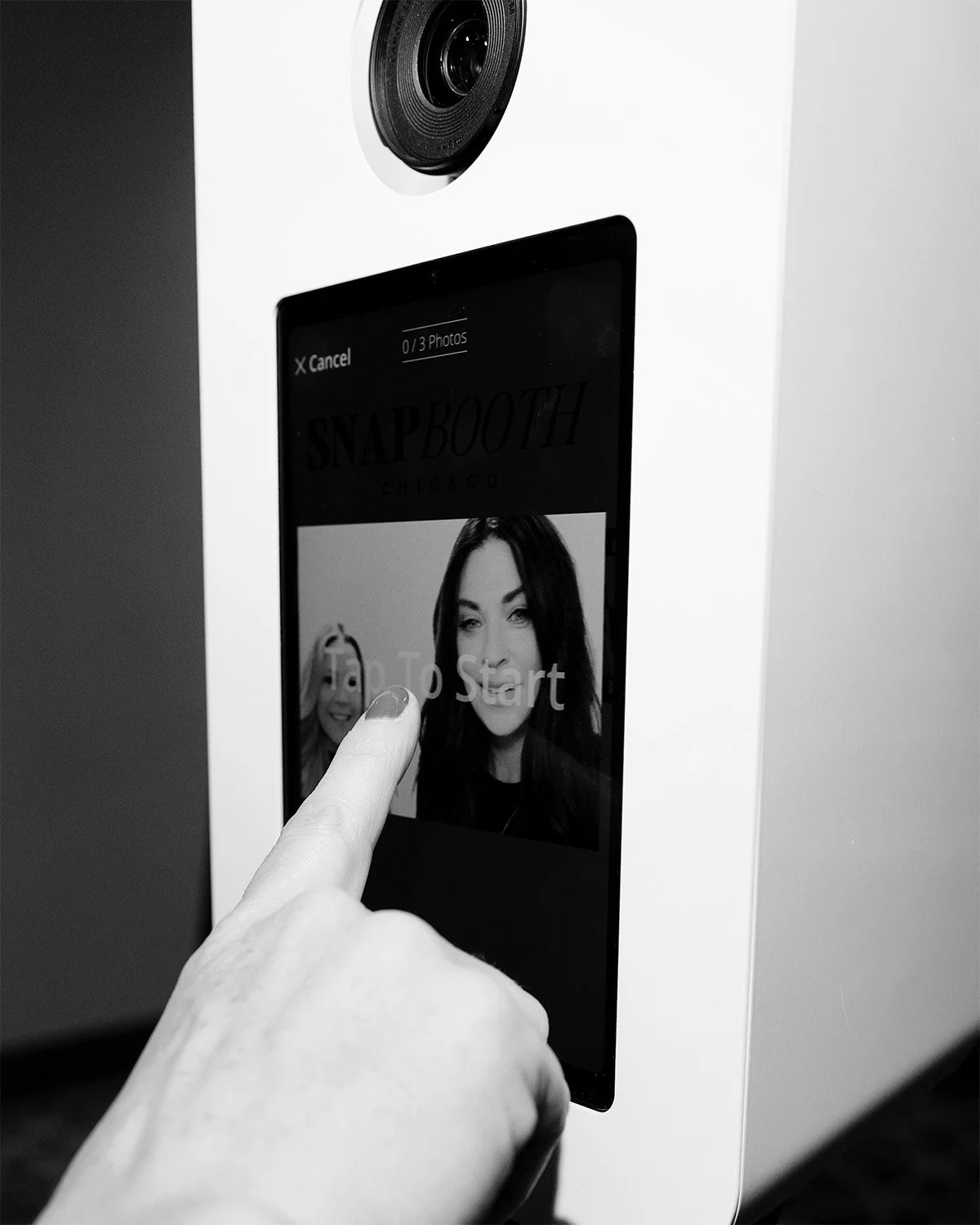 A person touching a touchscreen photo booth machine that displays a woman's face and the words 'Tap To Start'.