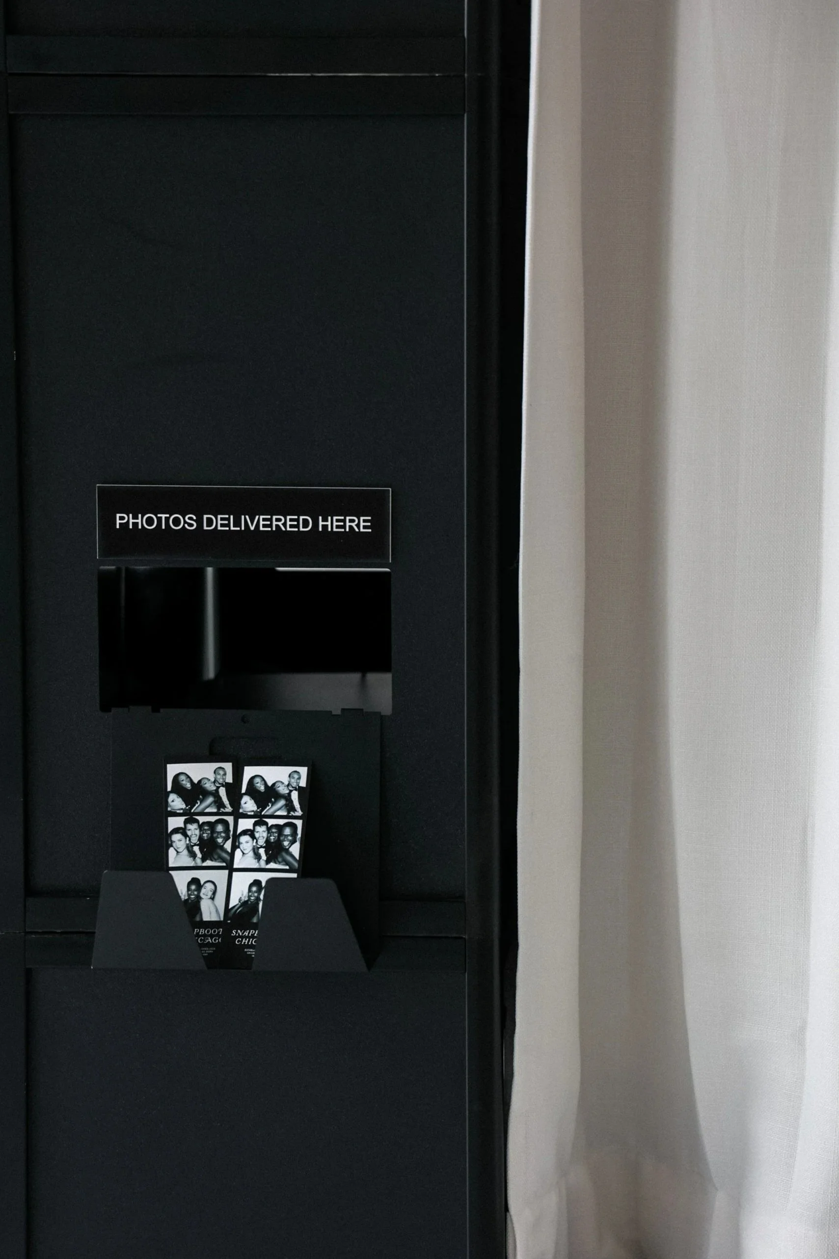 Photobooth with a sign that says 'Photos Delivered Here,' holding black-and-white photo strips with group pictures of people, next to a beige curtain.