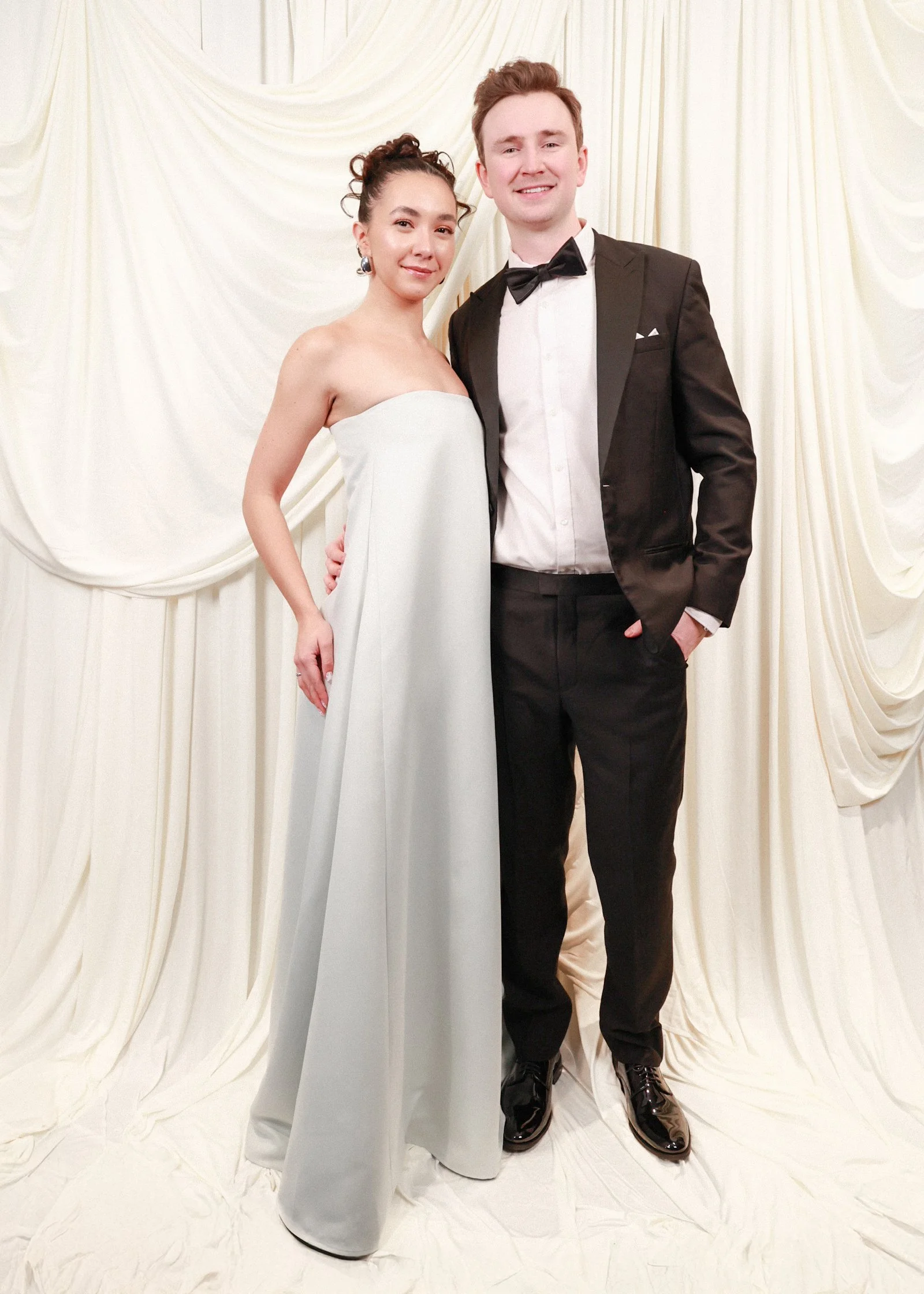 A formally dressed couple standing close together in front of a cream-colored draped fabric backdrop. The woman is wearing a strapless white gown, and the man is wearing a black tuxedo with a bow tie, smiling at the camera.