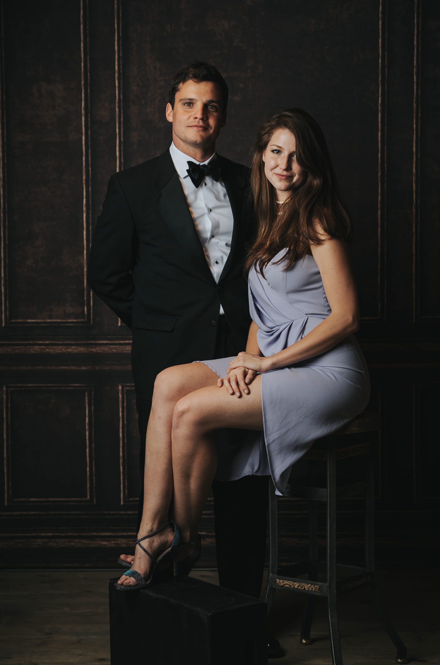 A man in a black tuxedo and a woman in a lavender dress pose against a dark wooden paneled background. The woman is sitting on a chair with her legs crossed and hands clasped on her lap, while the man stands behind her with one hand in his pocket.