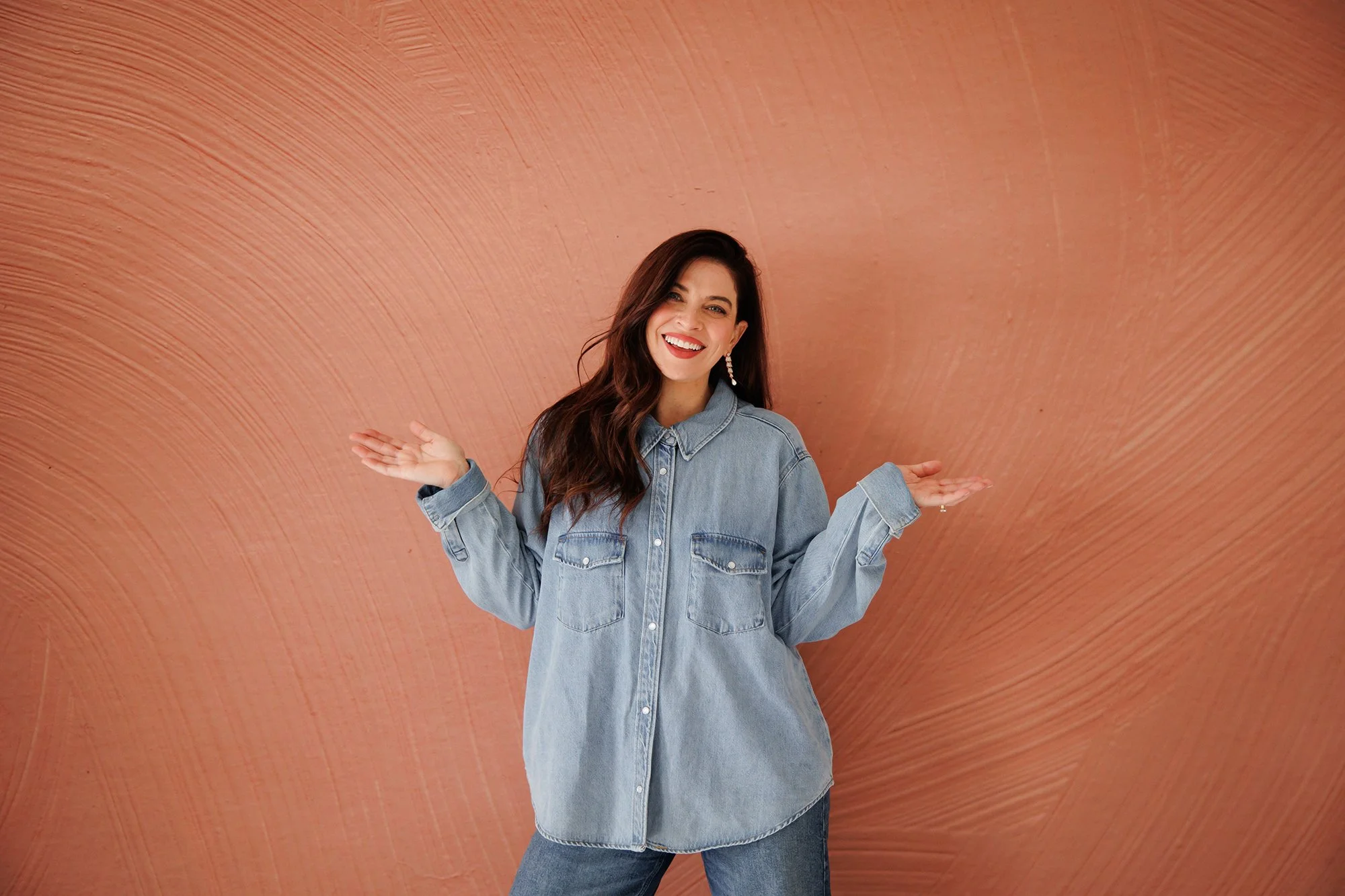 A woman smiling and gesturing with open hands in front of an orange, textured background, wearing a denim jacket and jeans.