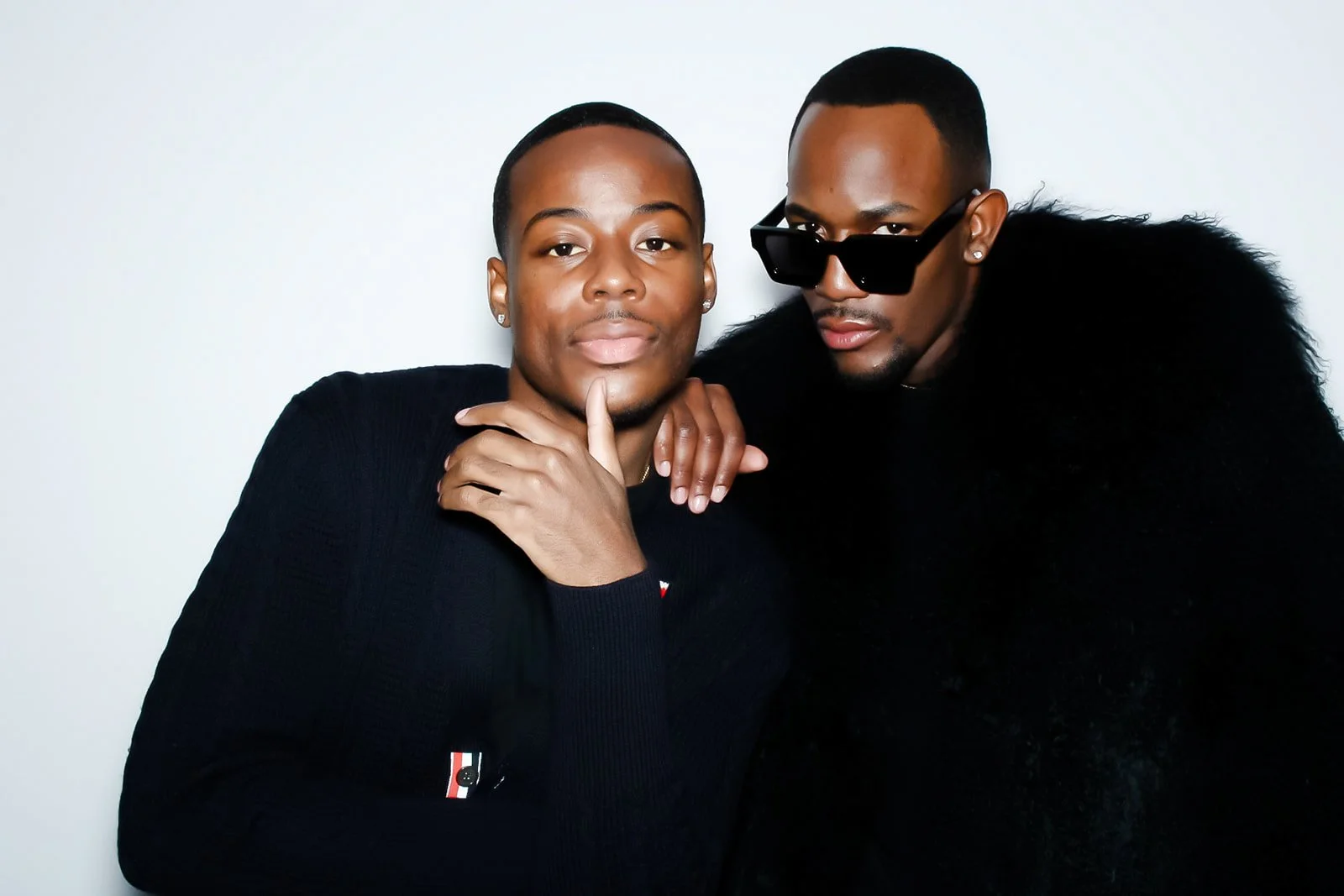 Two men posing closely together against a plain white background. The man on the left has a short haircut, is wearing a black top, and has a thoughtful expression with his hand near his chin. The man on the right has a similar haircut, wears black sunglasses and a black fluffy jacket, and is looking intently at the camera.