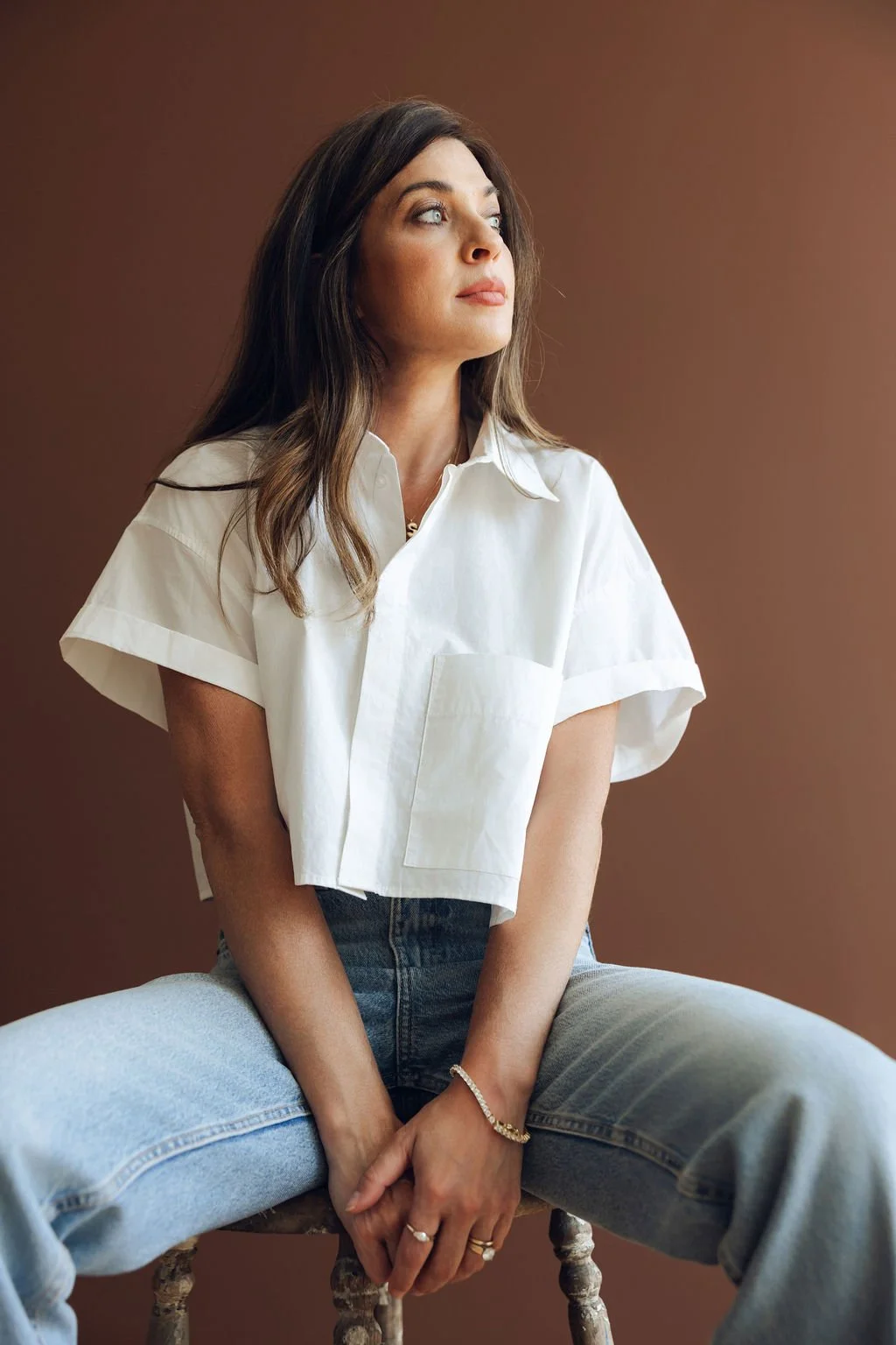 A woman with brown hair, fair skin, and blue eyes sits on a rustic wooden stool against a brown background. She is wearing a white short-sleeved button-up shirt, light blue jeans, and jewelry including rings, a bracelet, and a necklace. She looks thoughtfully to her left.